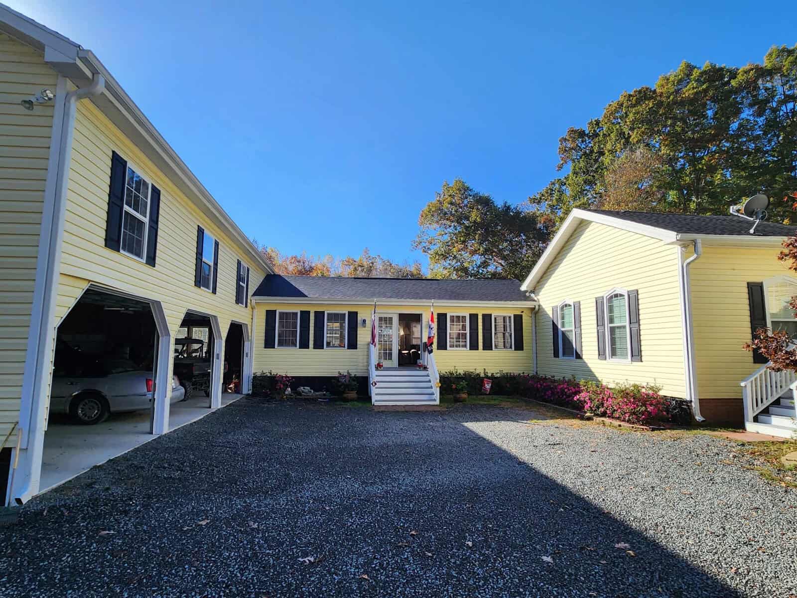 A yellow house with white trim is pictured under a clear blue sky. It has a central entrance with steps and a driveway leading to a garage on the left. The yard is landscaped with small shrubs and trees.