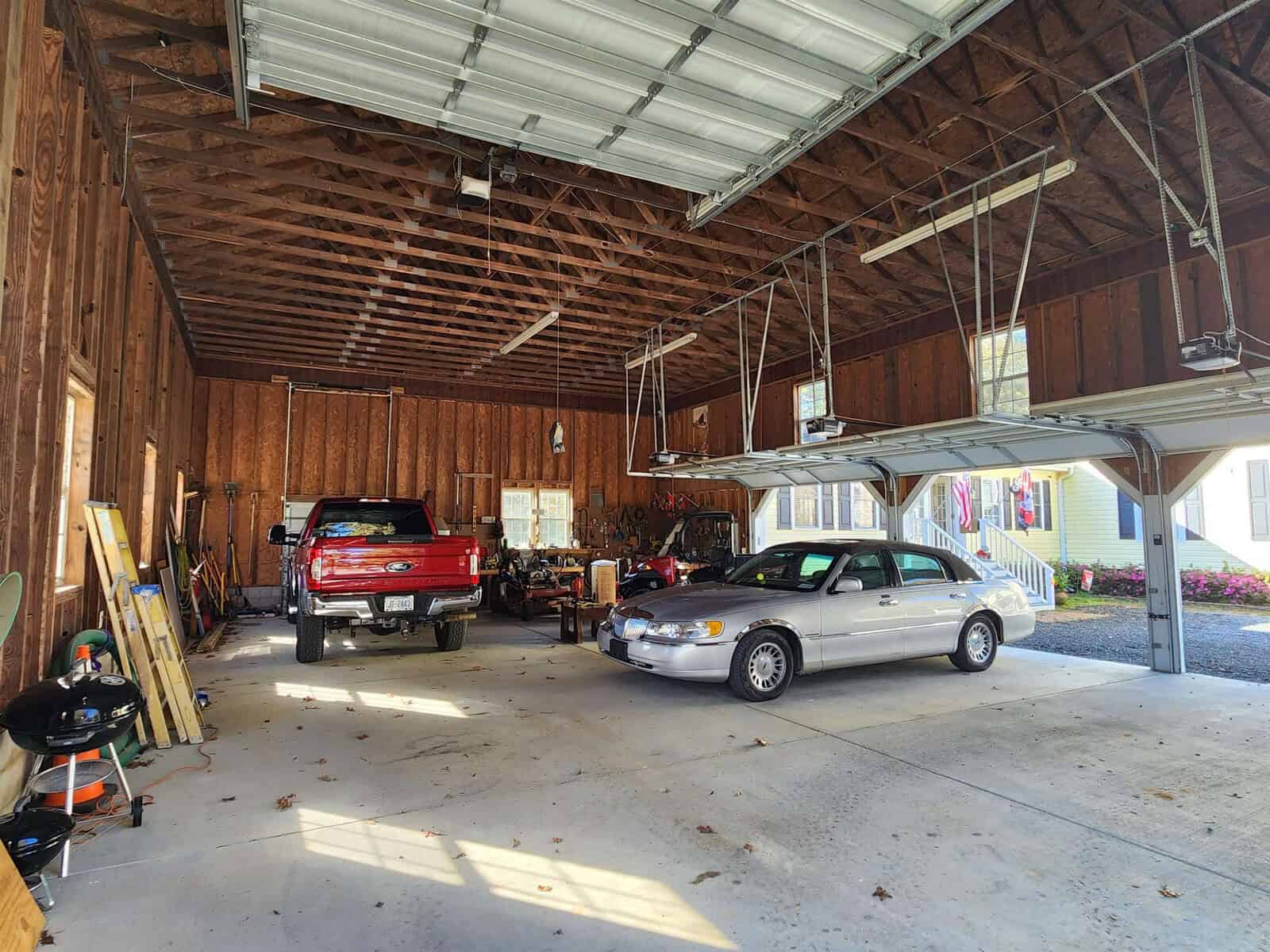 Interior view of a spacious garage with wooden walls and an open ceiling. A red truck and a silver car are parked inside. Shelves and various items are stored along the walls, and a second garage door is partially visible.