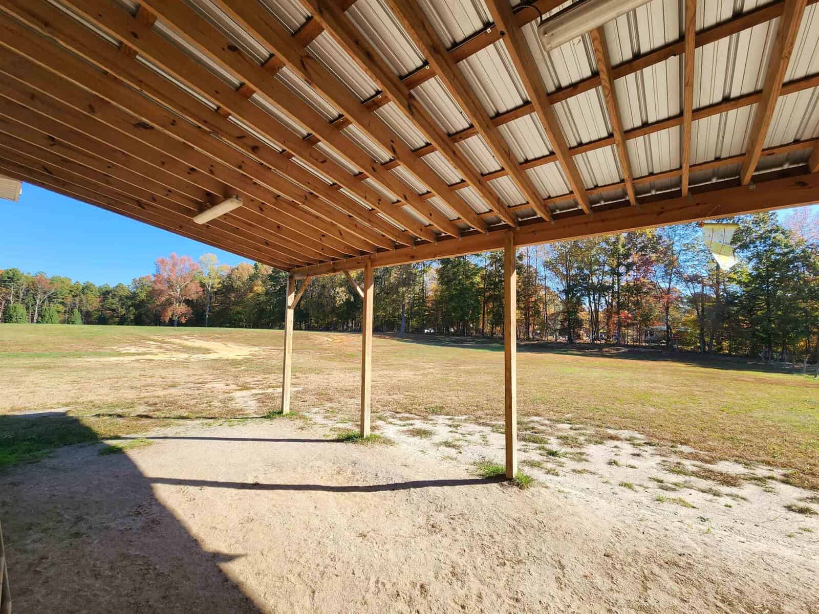 A wooden pavilion with a metal roof overlooks an open grassy field bordered by trees with autumn foliage. The ground under the pavilion is sandy, and the sky is clear and blue.