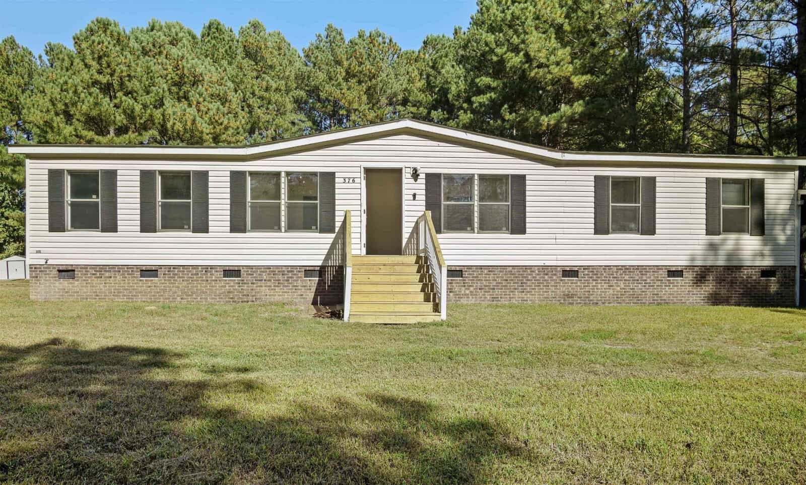 A single-story manufactured home with beige siding and a small front porch is set amidst a grassy yard with trees in the background, located at 376 Huff Road, Henderson NC.
