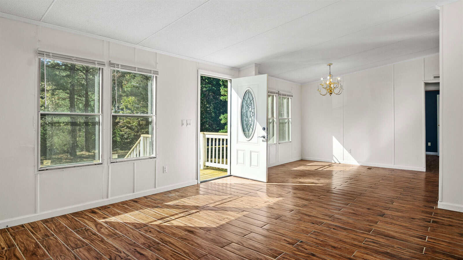 Empty living room with hardwood floors, white walls, multiple windows, a glass-paneled door leading to a small balcony, and a chandelier.