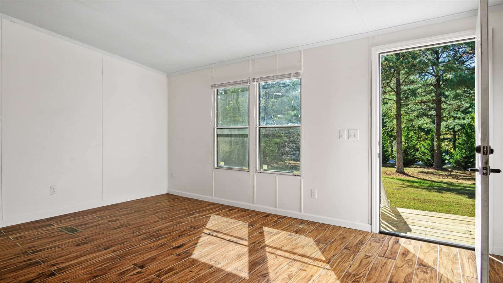 An empty room with wooden parquet flooring, white walls, and large windows providing a view of trees outside, with a door leading to an exterior wooden deck.