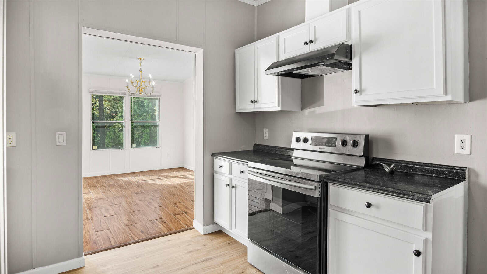 A clean and unoccupied kitchen with white cabinetry, black countertops, and stainless steel appliances adjacent to a room with a chandelier and a view of a window.