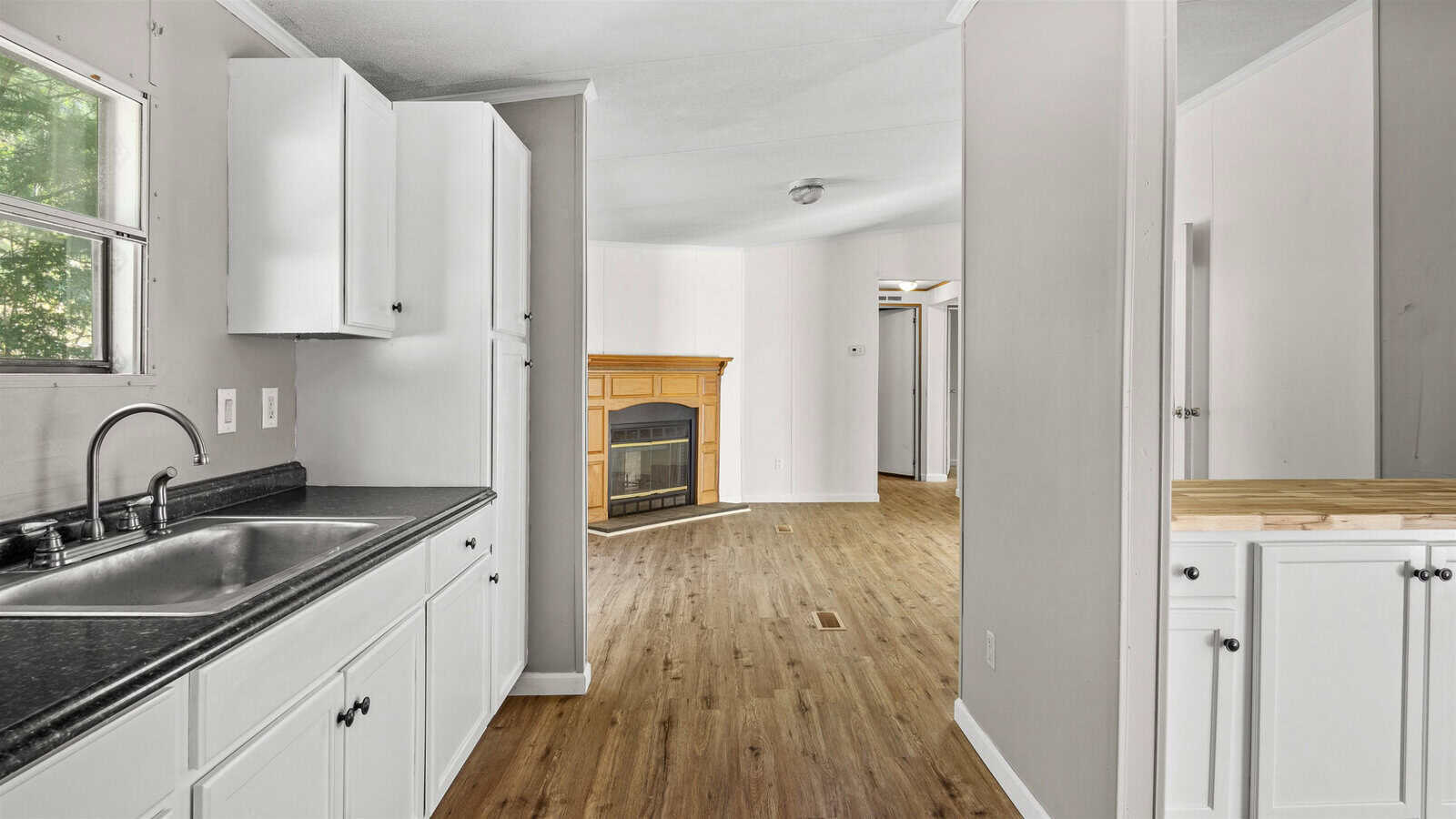 A view into an empty, renovated kitchen with white cabinetry and contrasting dark countertops, adjacent to a living area with a fireplace and hardwood flooring, suggesting a modern interior space.