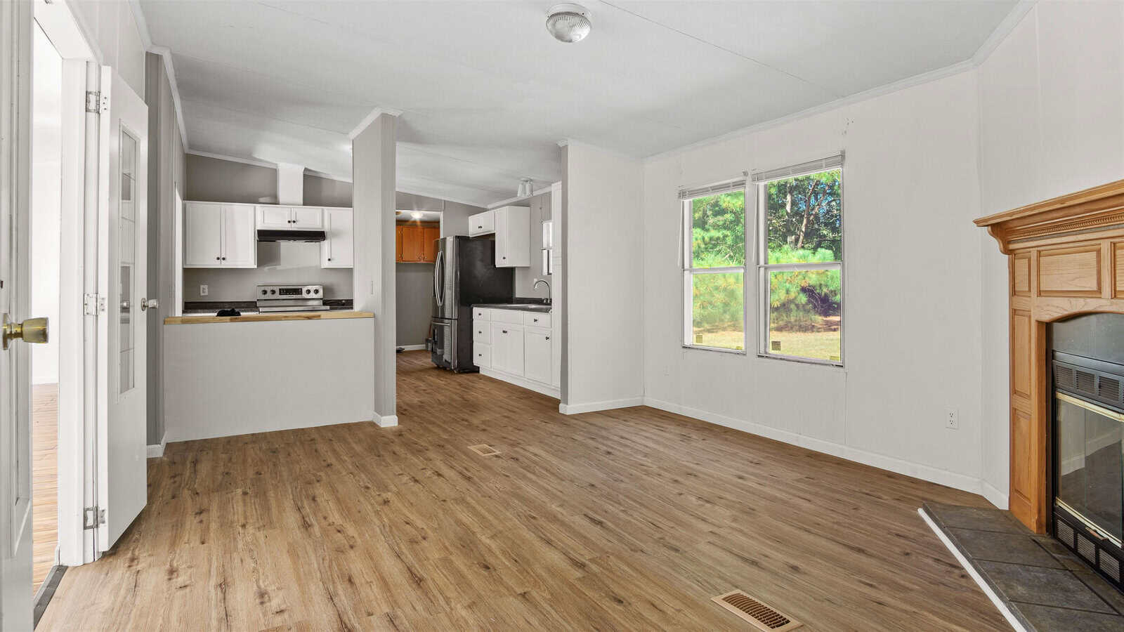 A bright, empty room featuring an open-plan kitchen with modern appliances and light wooden flooring, leading to a living area with a fireplace and windows overlooking greenery.