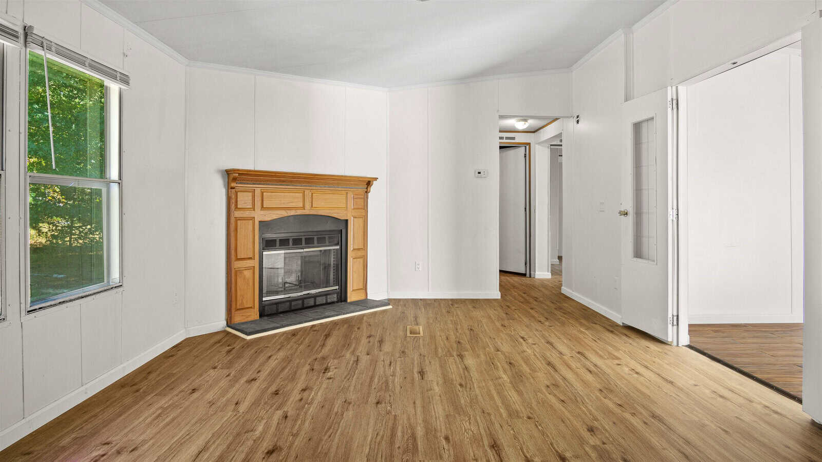 Empty room with a hardwood floor, white walls, a fireplace with a wooden mantel, and a hallway in the background.