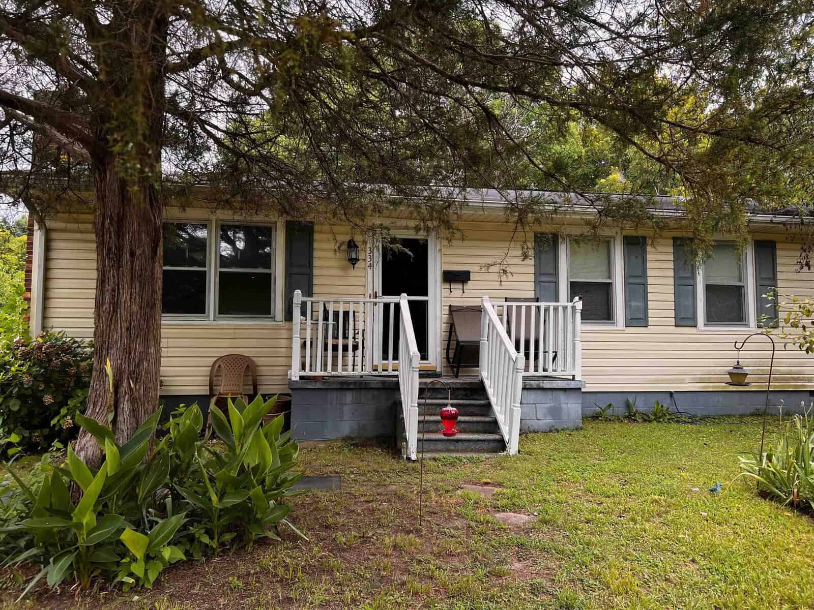 A single-story house at 334 Pearl Street, Henderson NC, with yellow siding and white trim, featuring a small front porch with stairs and a wooden railing. The house is partially obscured by greenery