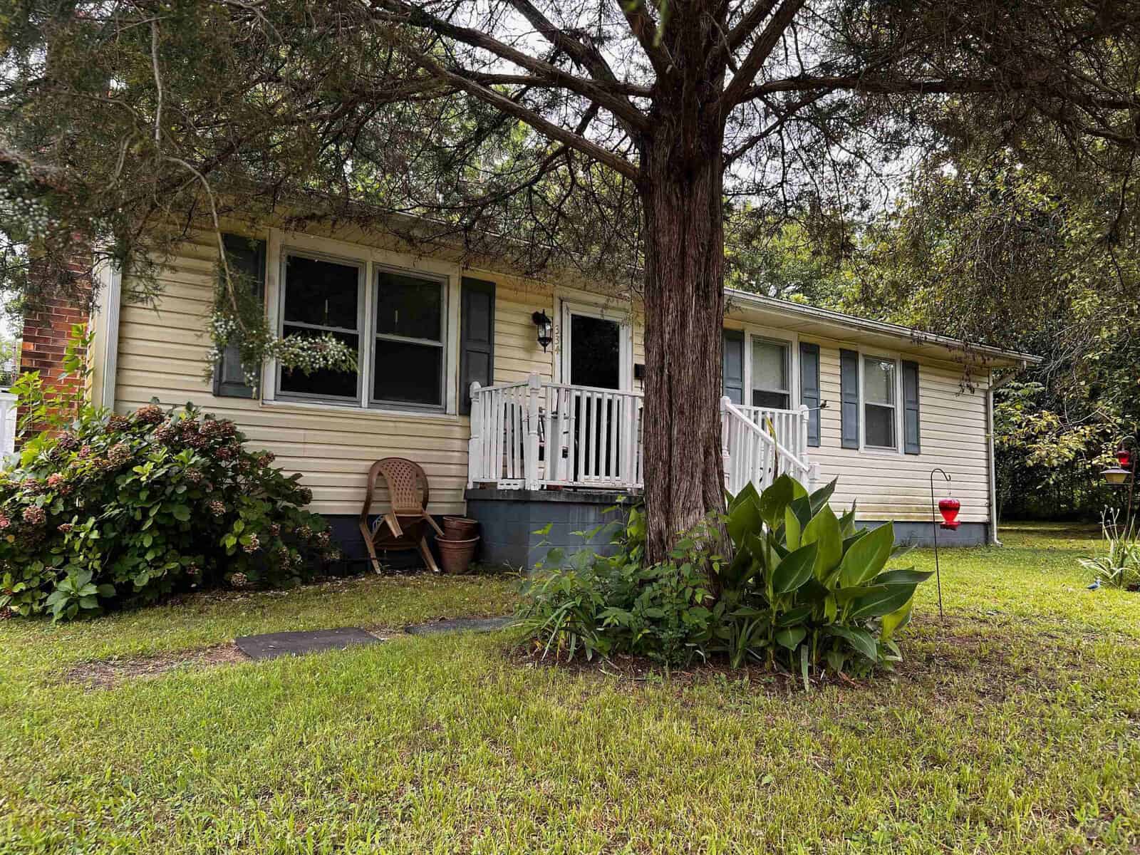 Single-story house with a front porch, surrounded by greenery and a large tree in the foreground.