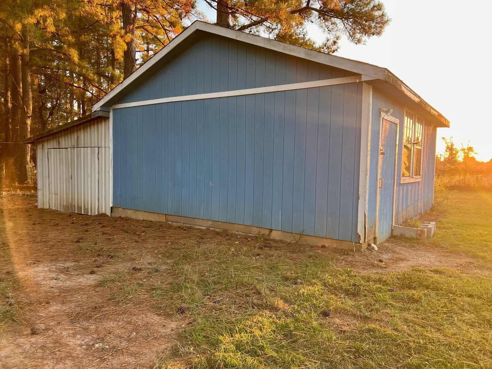 Blue wooden shed with a sloping roof situated in a field with trees in the background during sunset.