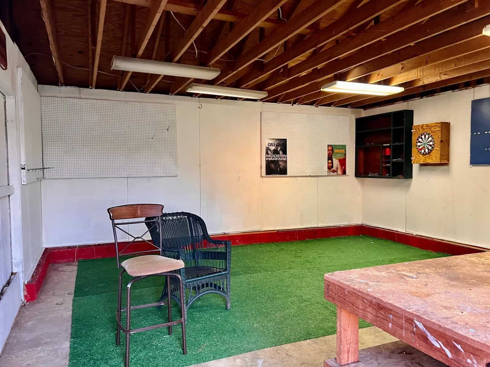 An interior space featuring a dart board on the wall, a folding chair and a black plastic chair on an artificial grass mat, with a wooden table in the foreground and exposed wooden beams above.