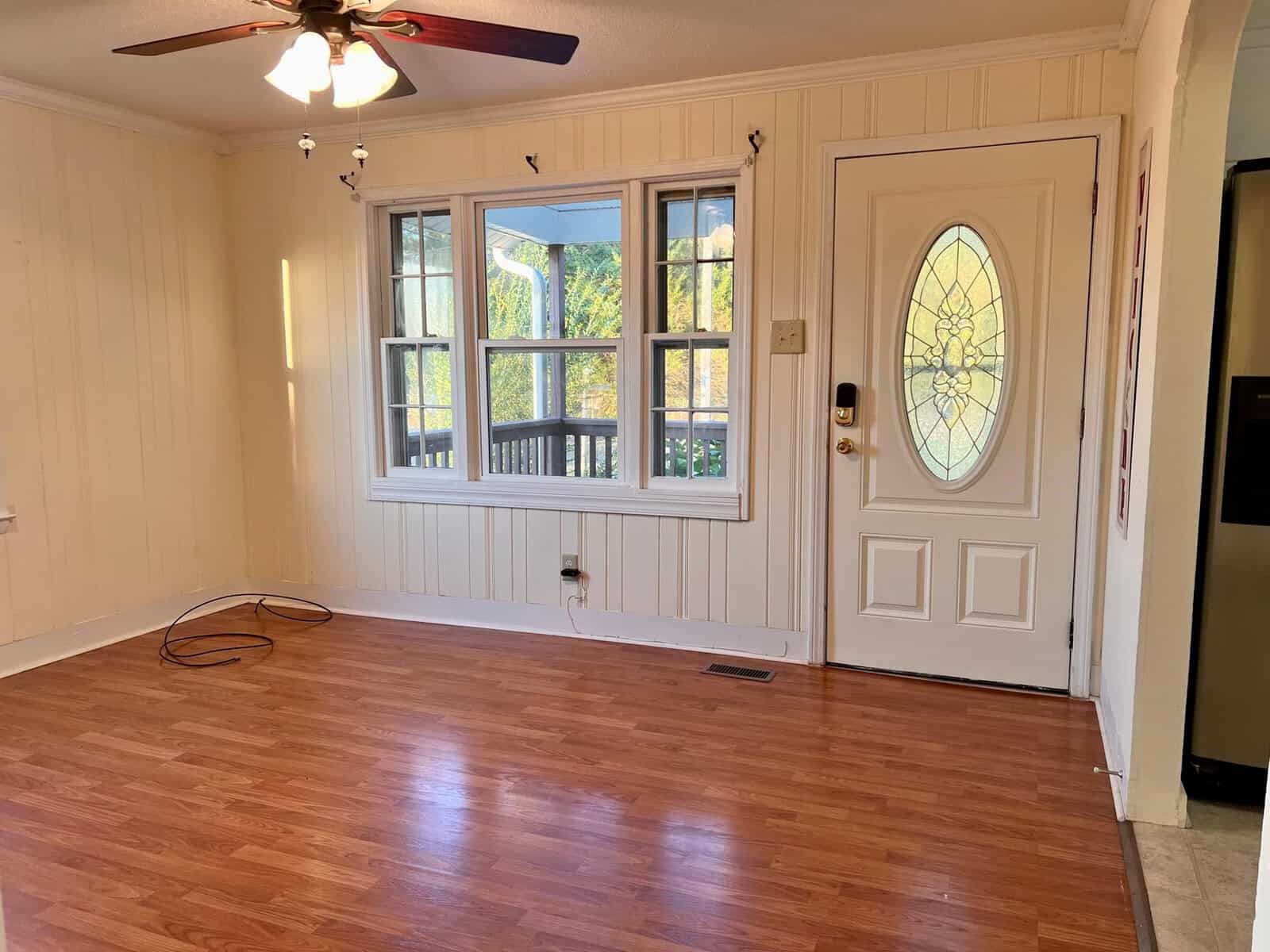 An empty room featuring wooden flooring, a white front door with decorative glass, flanked by a window looking out onto a balcony, and a ceiling fan above.