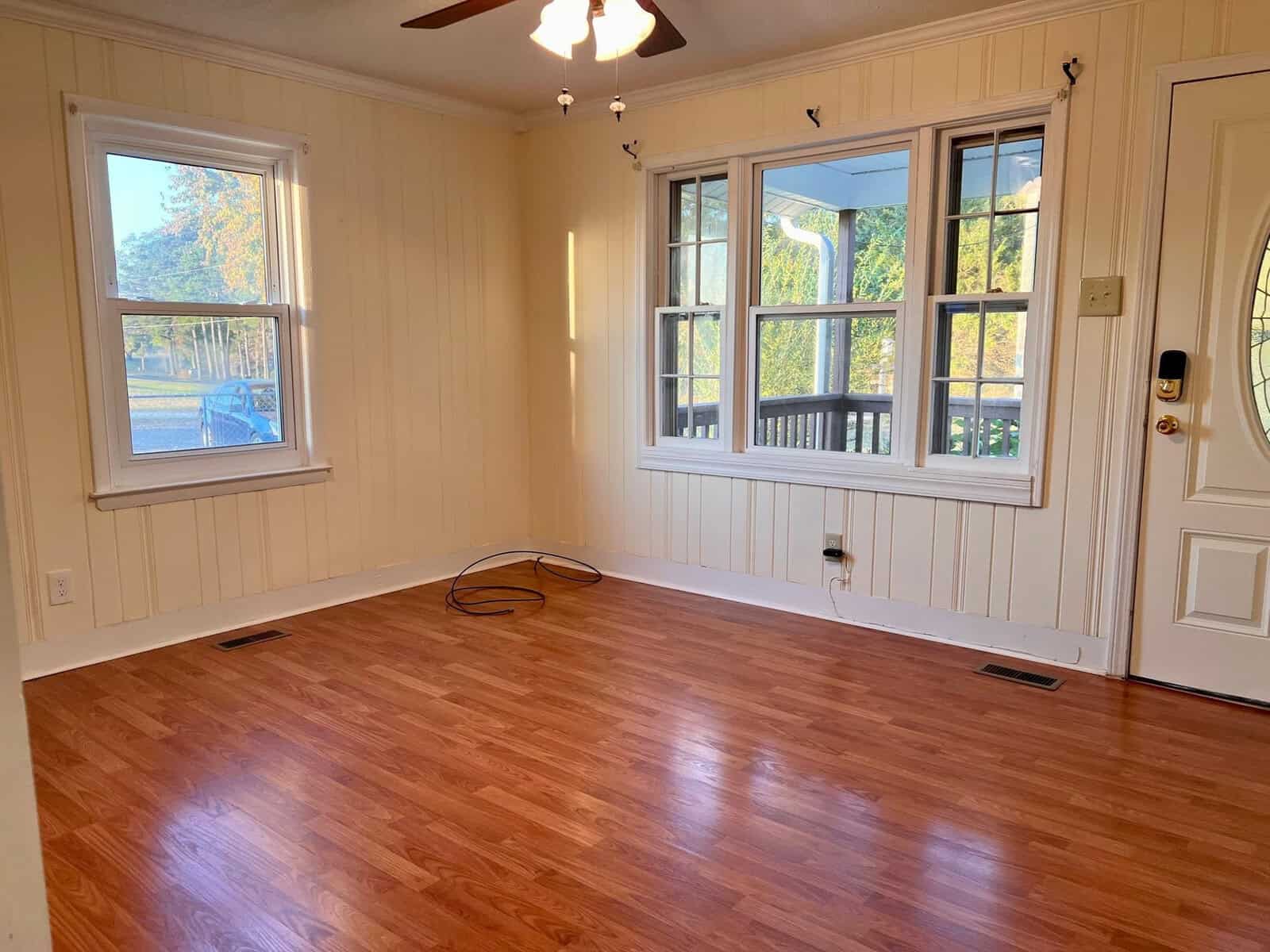An empty room with wooden flooring, white walls with vertical paneling, a chandelier, and two windows providing natural light. a door with a digital lock is visible on the right.