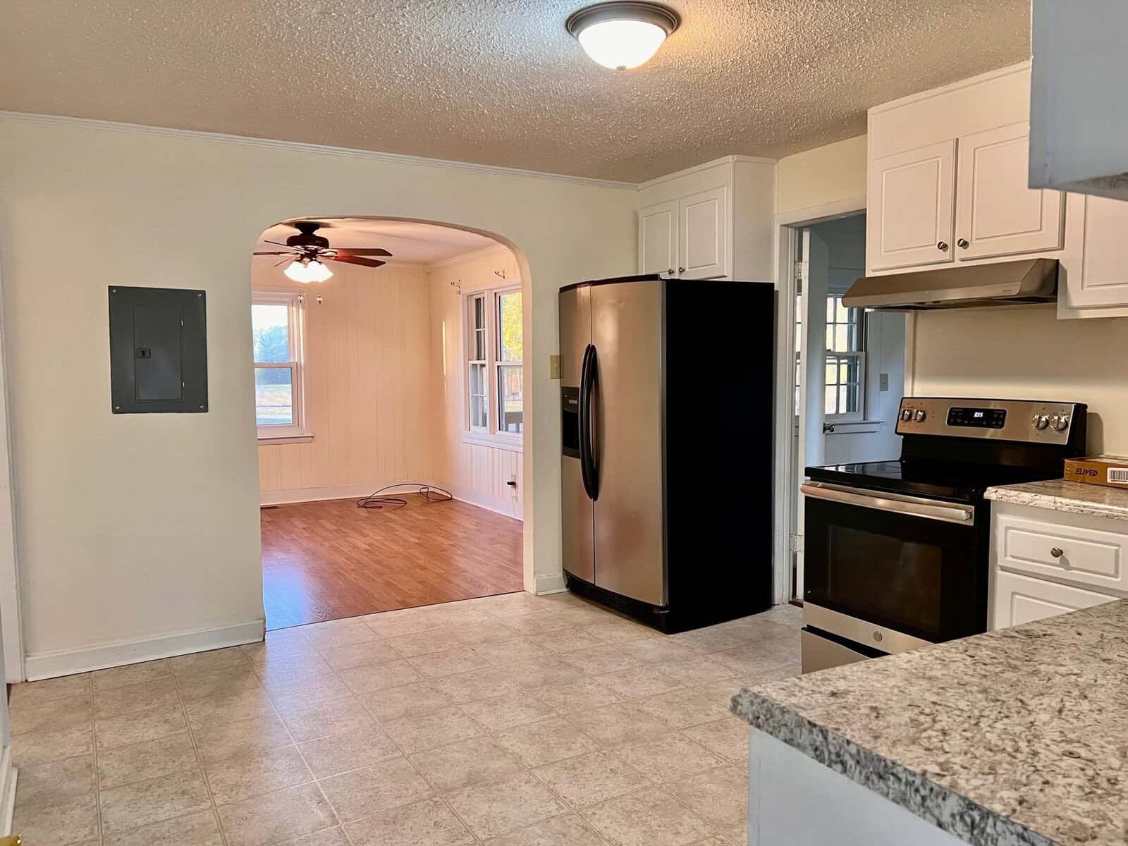An empty kitchen with white cabinets, black appliances, and a tiled floor, looking into an adjoining room with wooden flooring.