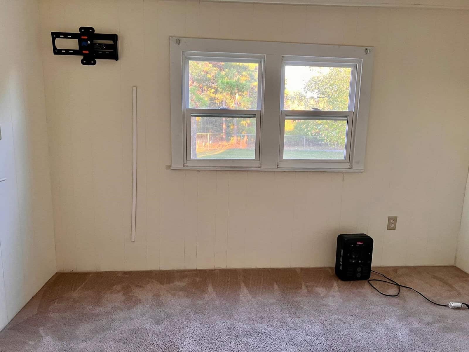 An empty room with cream-colored walls and carpet, featuring a double window with a view of trees, a thermostat on the left wall, and a small black heater on the floor near an electrical outlet.