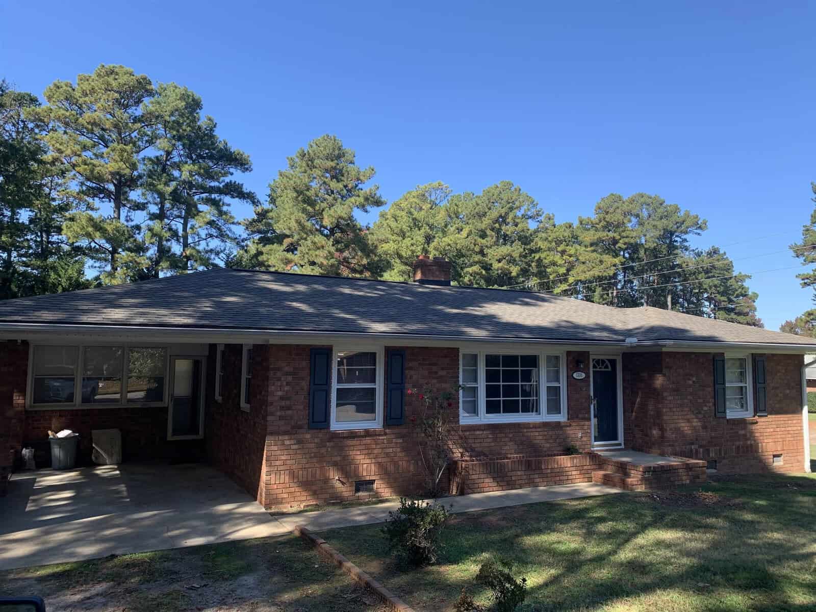 A single-story brick house with a black shingled roof, featuring a covered porch, situated under a clear blue sky with pine trees in the background.
