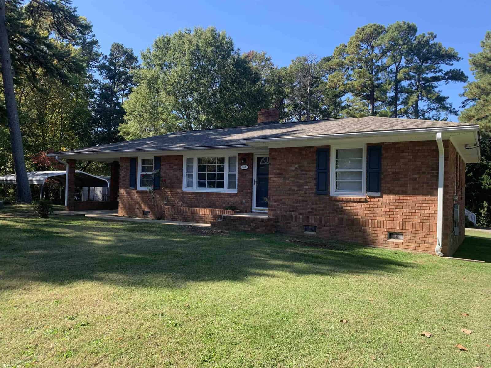 A single-story brick house with a white door and window frames, featuring a carport on the left and surrounded by a grassy lawn with trees in the background.