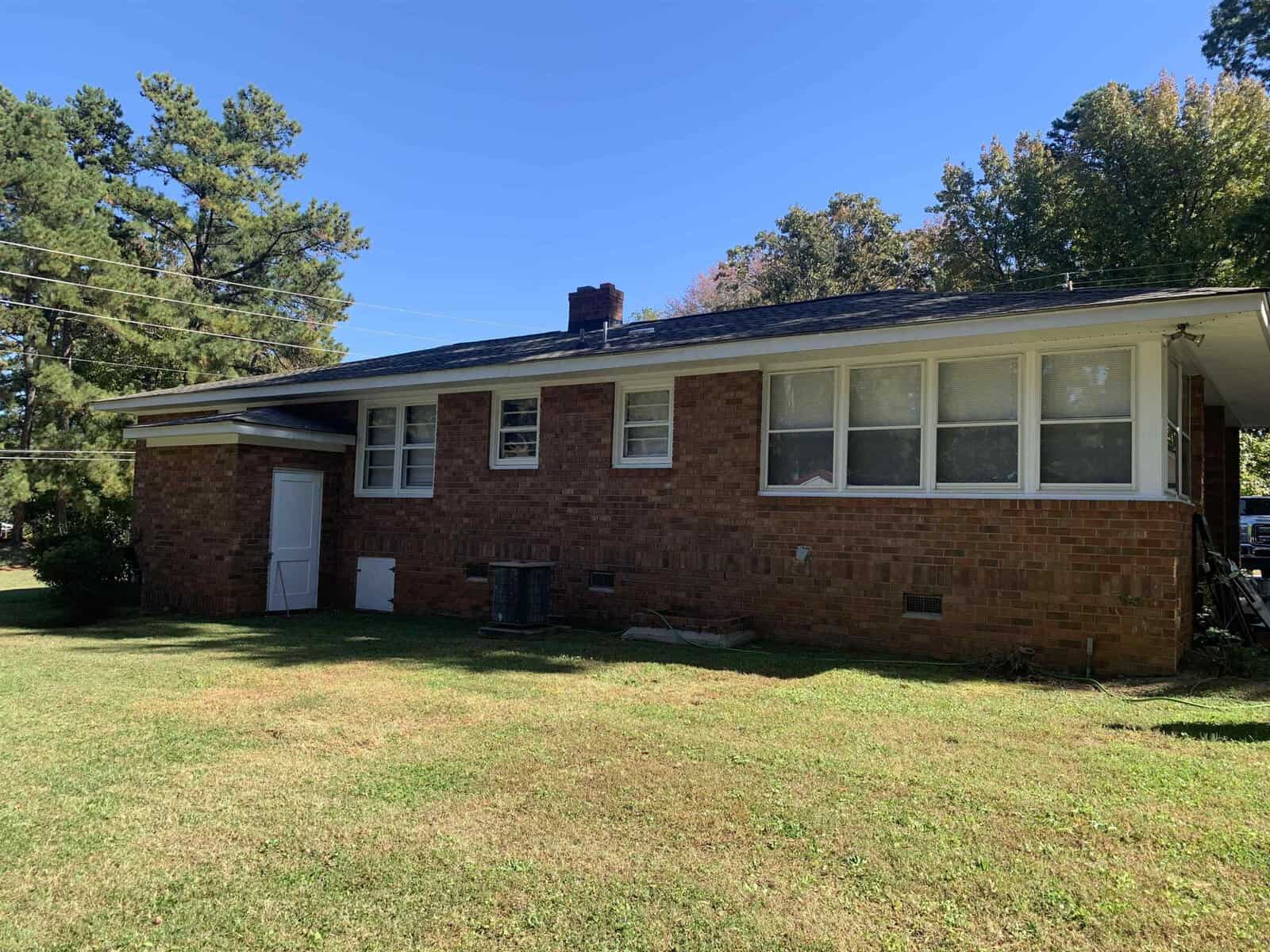 A single-story brick house with a white door and window frames, featuring a chimney and front lawn on a sunny day.