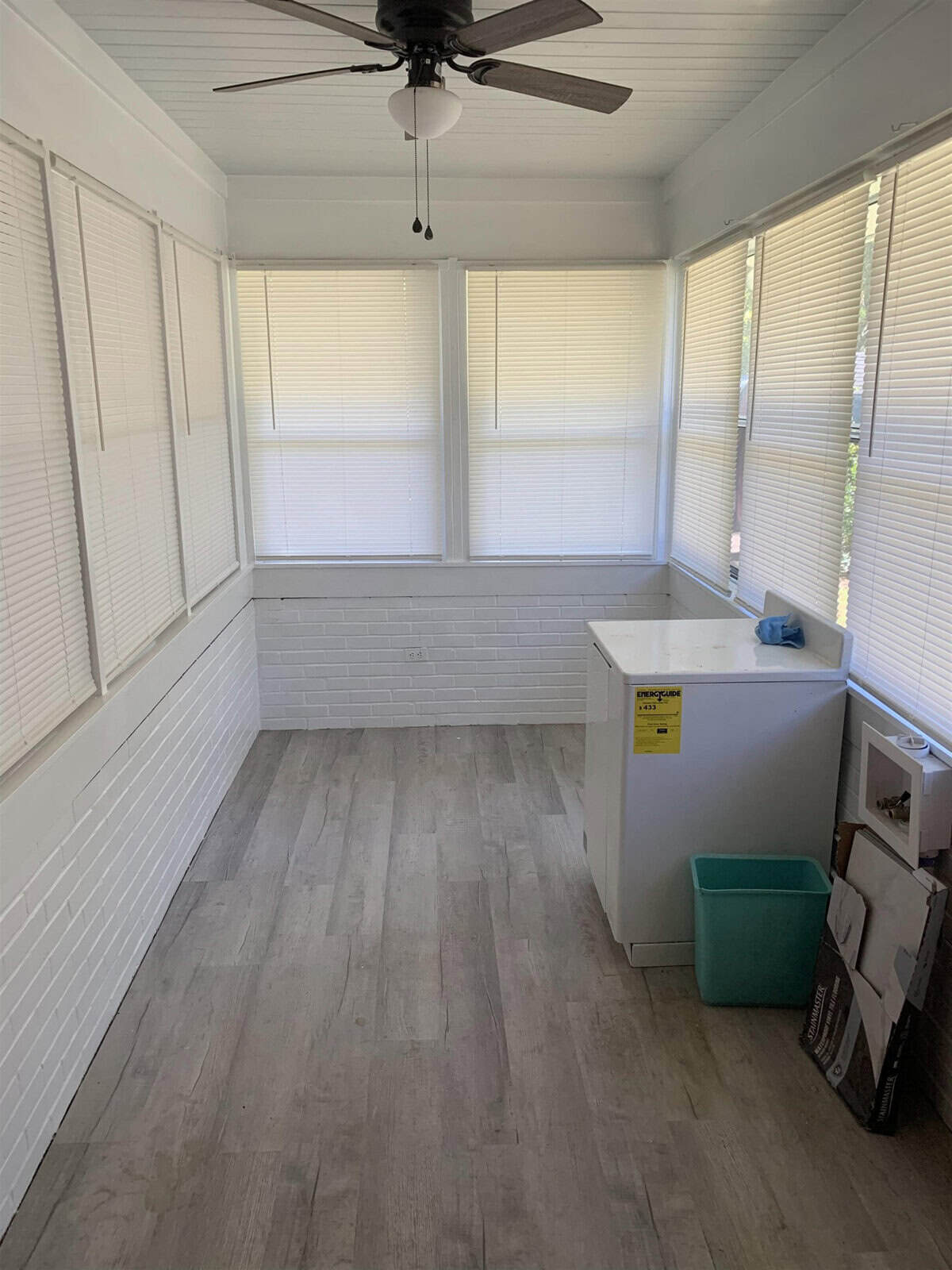 A clean, empty room with white walls and a faux wood floor, featuring a ceiling fan, multiple windows with white blinds, and a small white counter with a backsplash on one side. a blue recycling bin and a green container are situated in the corner.