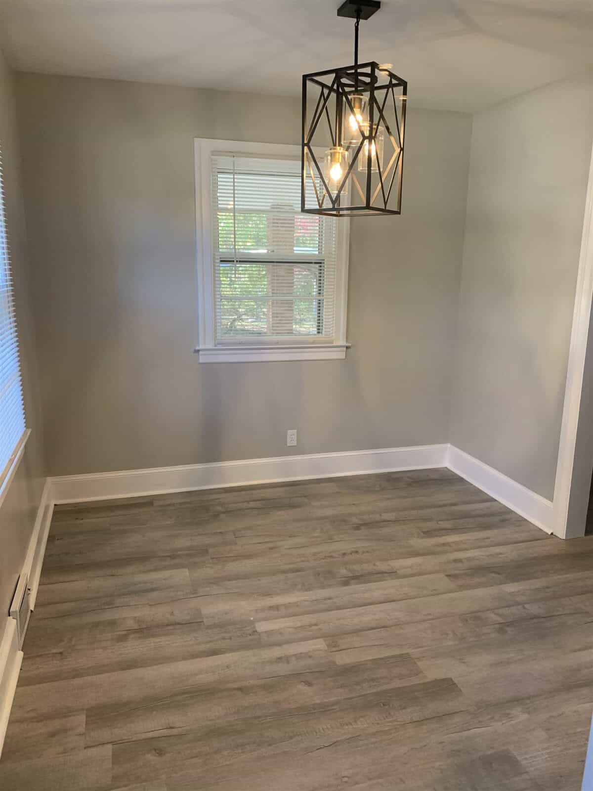 An empty room with gray flooring, taupe walls, a window, and a black geometrical chandelier with exposed bulbs.