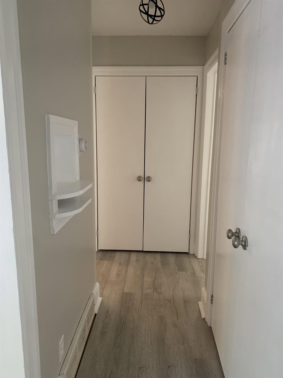 A hallway featuring a closed double door at the end, with hardwood flooring, a small shelf on the left wall, and doors on the right.