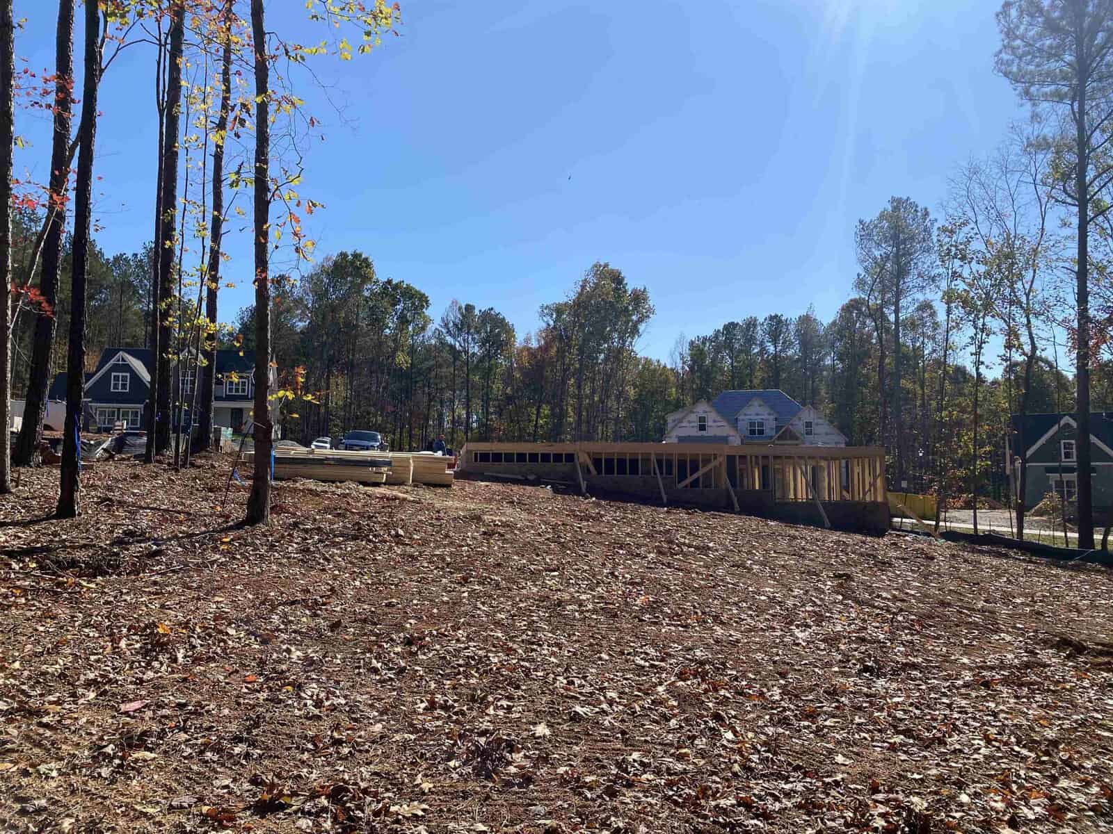 A construction site in a wooded area with partially built wooden structures. Trees with autumn leaves surround the site. In the background, there are several completed houses. The sky is clear and sunny.