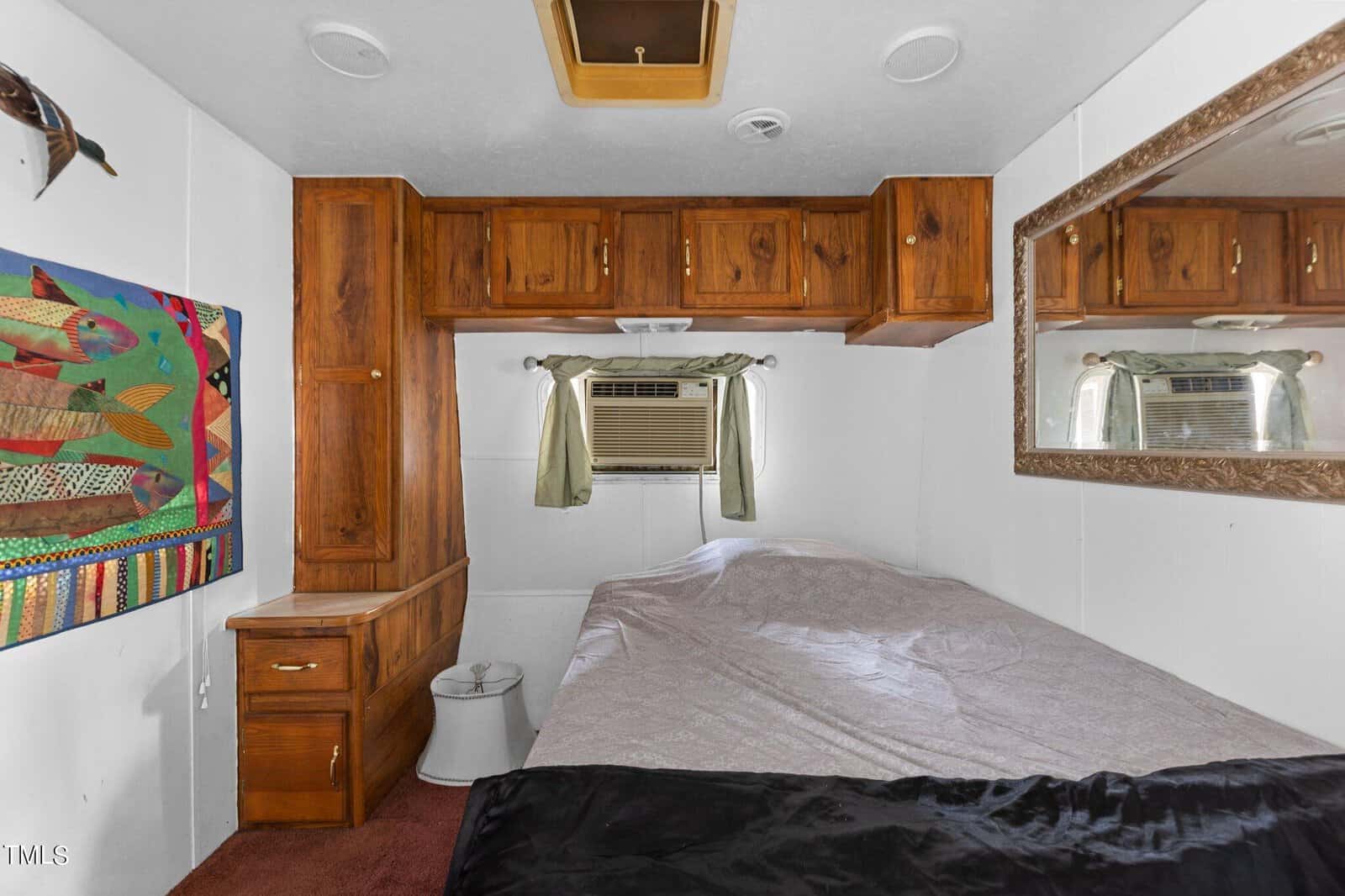 Small bedroom with wood cabinets, a bed, a window with an air conditioner, a large wall mirror, and a colorful fish-themed wall hanging. The room has white walls and a red carpet floor.