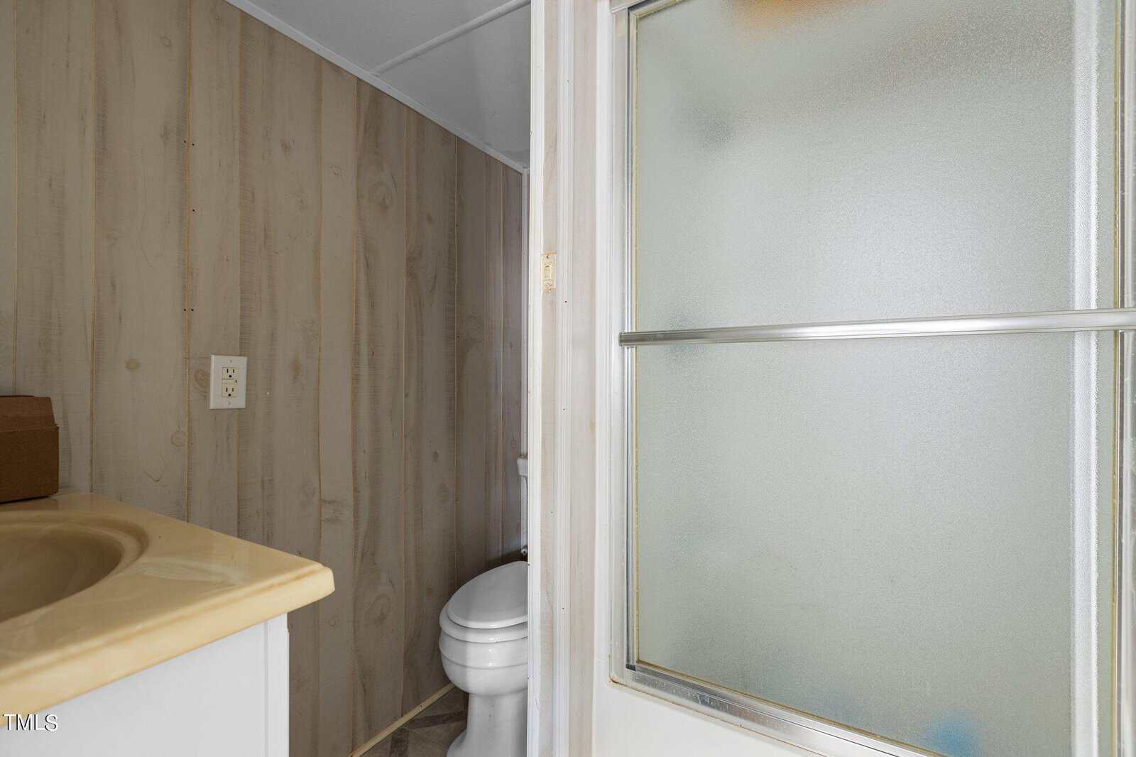 Small bathroom with wood-patterned walls, a beige countertop with sink, a toilet, an electrical outlet, and a frosted glass sliding shower door partially visible on the right.