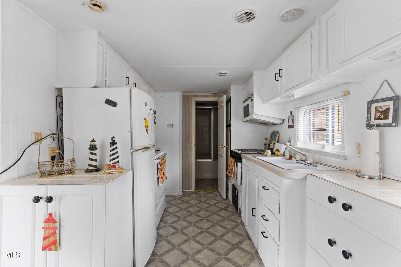 A small, white kitchen with cabinets, a stove, a fridge, and a microwave. Nautical decor items, like lighthouse figurines, are on the counters. A hallway leads to a bathroom at the back. Natural light enters through a window.