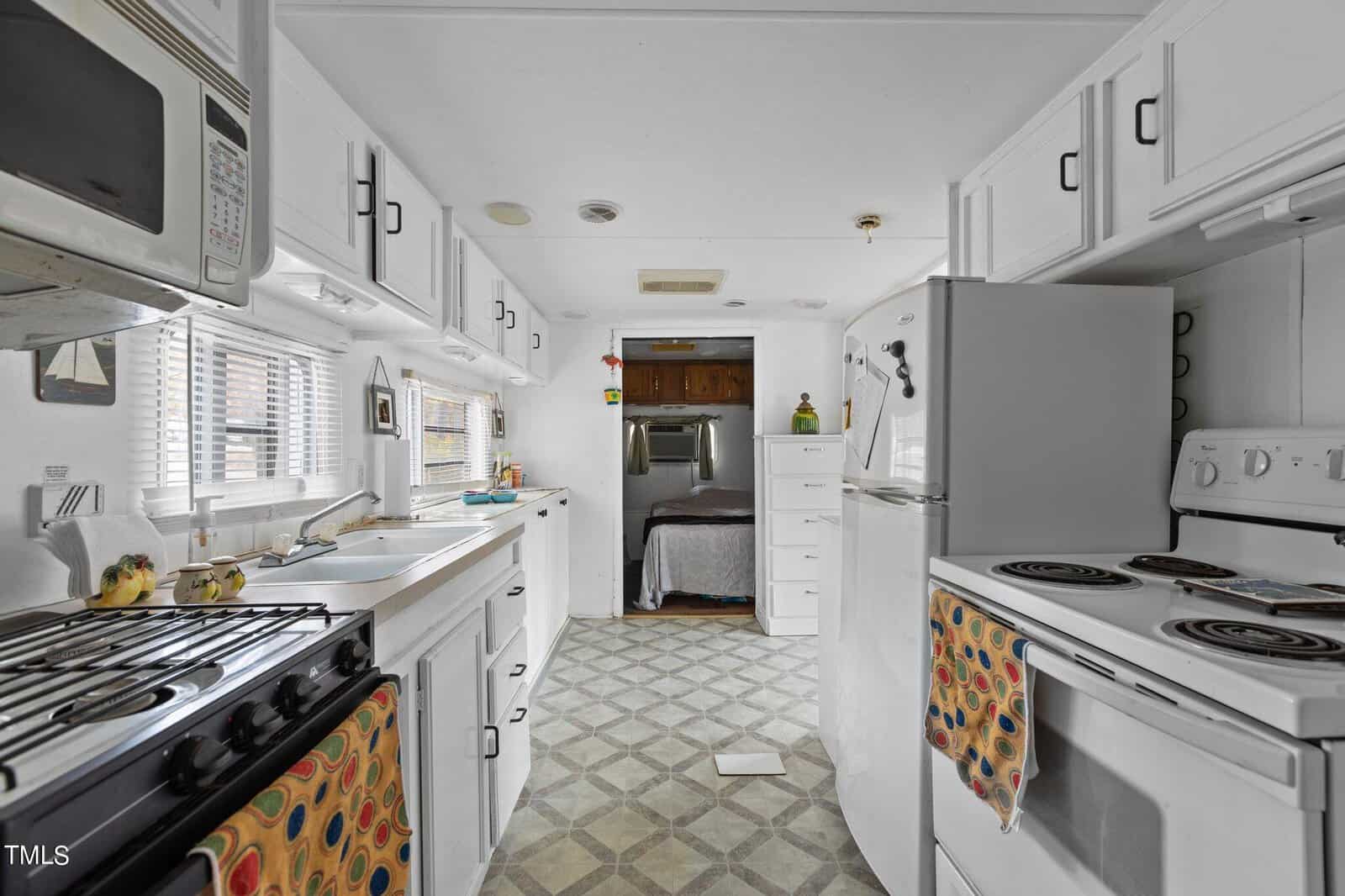A narrow, white kitchen with white cabinets and appliances, patterned towels, a window above the sink, and a view into a bedroom at the far end. The floor has a gray geometric pattern.
