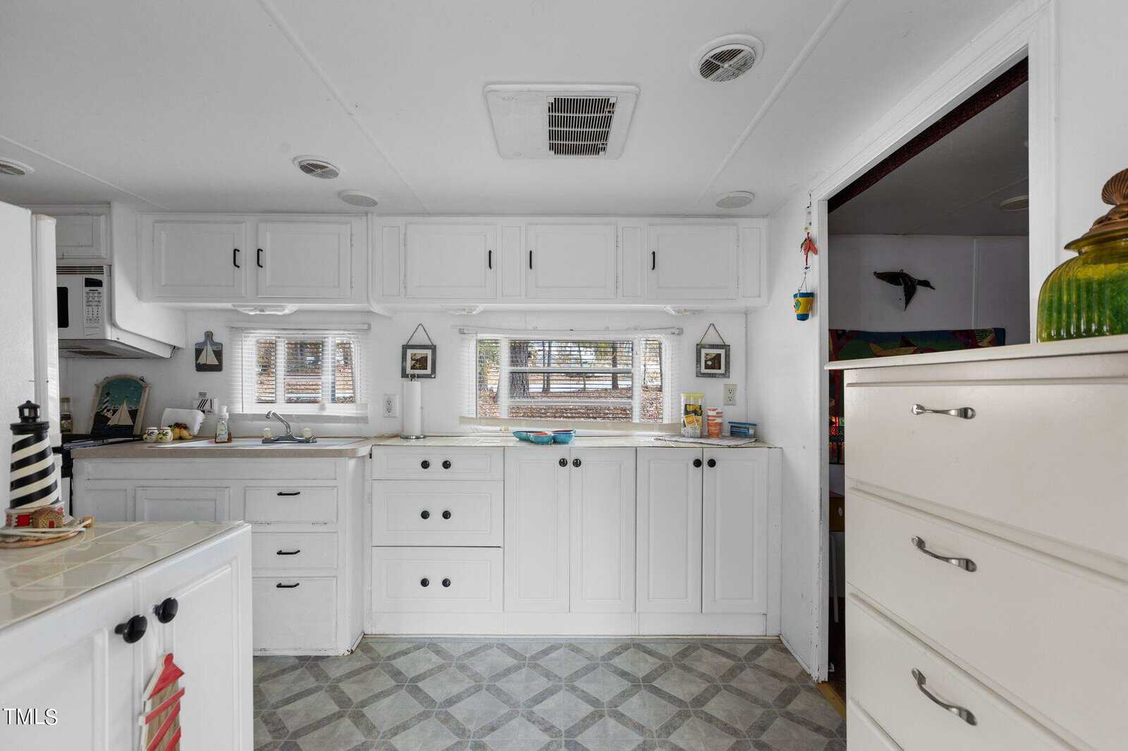 A small, white kitchen with upper and lower cabinets, double windows, and patterned floor tiles. Various kitchen items are on the countertops, and a white dresser stands to the right.