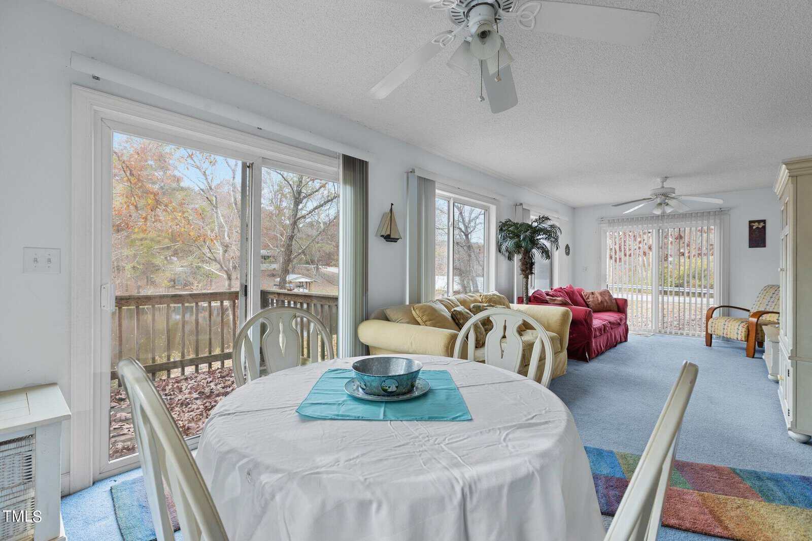 Bright living and dining area with large windows and sliding glass doors, featuring a round table with white chairs, cozy sofas, a colorful rug, and views of trees and a wooden deck outside.