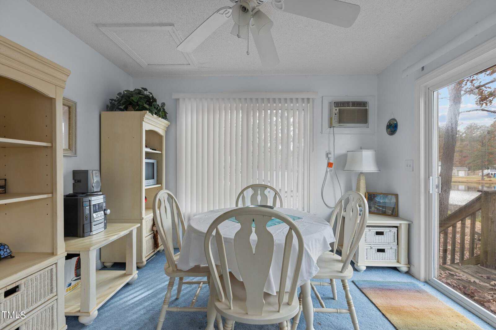 A bright dining area with a round table and four chairs, surrounded by light-colored furniture. Large windows and sliding glass doors let in natural light, with a view of trees and a lake outside.