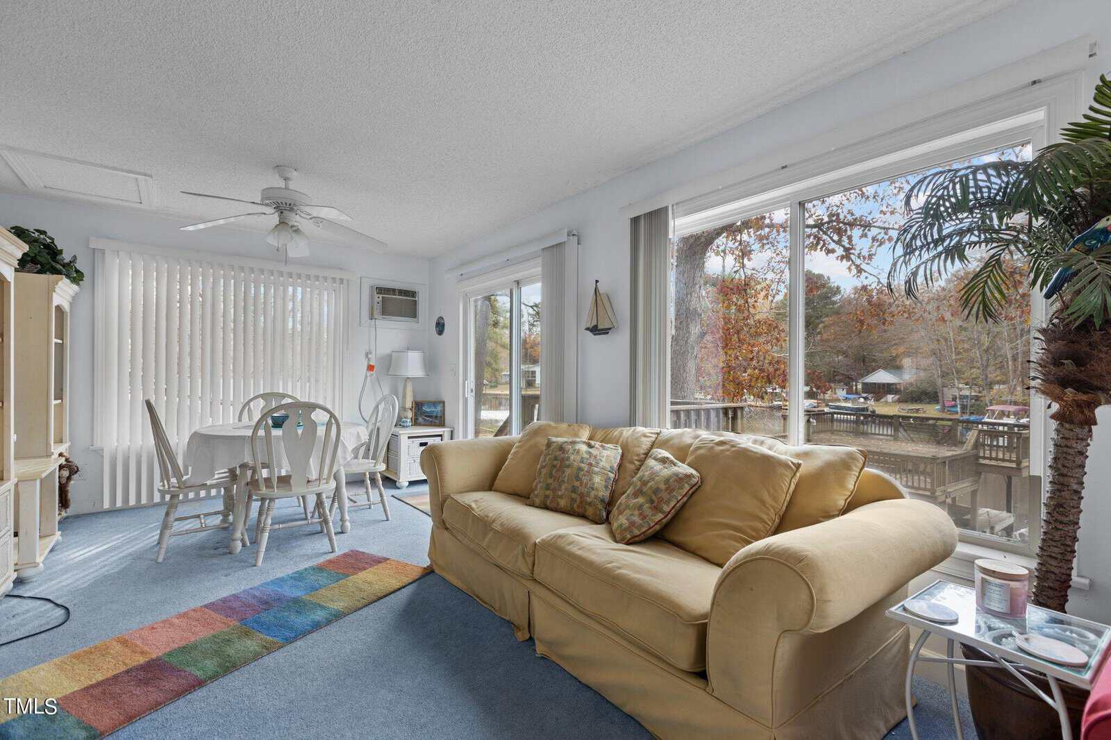 Bright sunroom with large windows, a yellow sofa with pillows, a small white dining table with chairs, and a colorful rug. Outside, trees and a wooden deck are visible. A palm plant sits by the window.