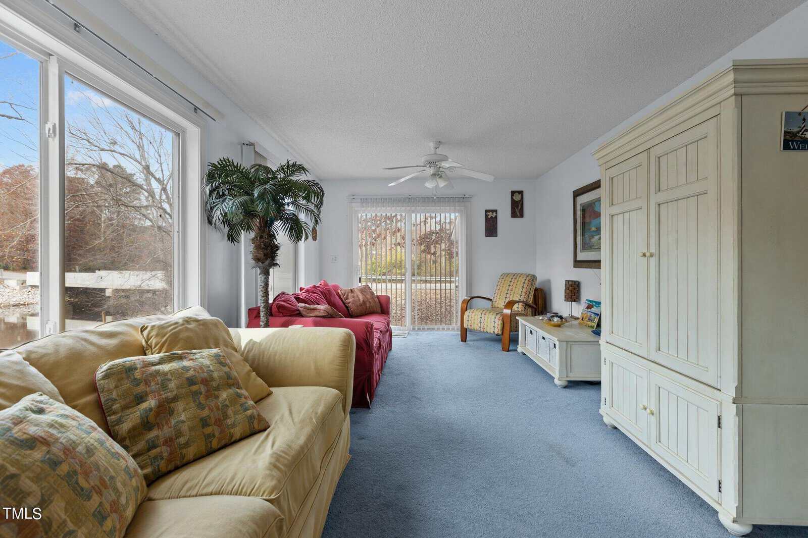 A bright living room with a large window, sliding glass doors, beige sofa, red loveseat, patterned armchair, white entertainment center, potted palm, and blue carpet. Artwork and throw pillows add cozy touches.