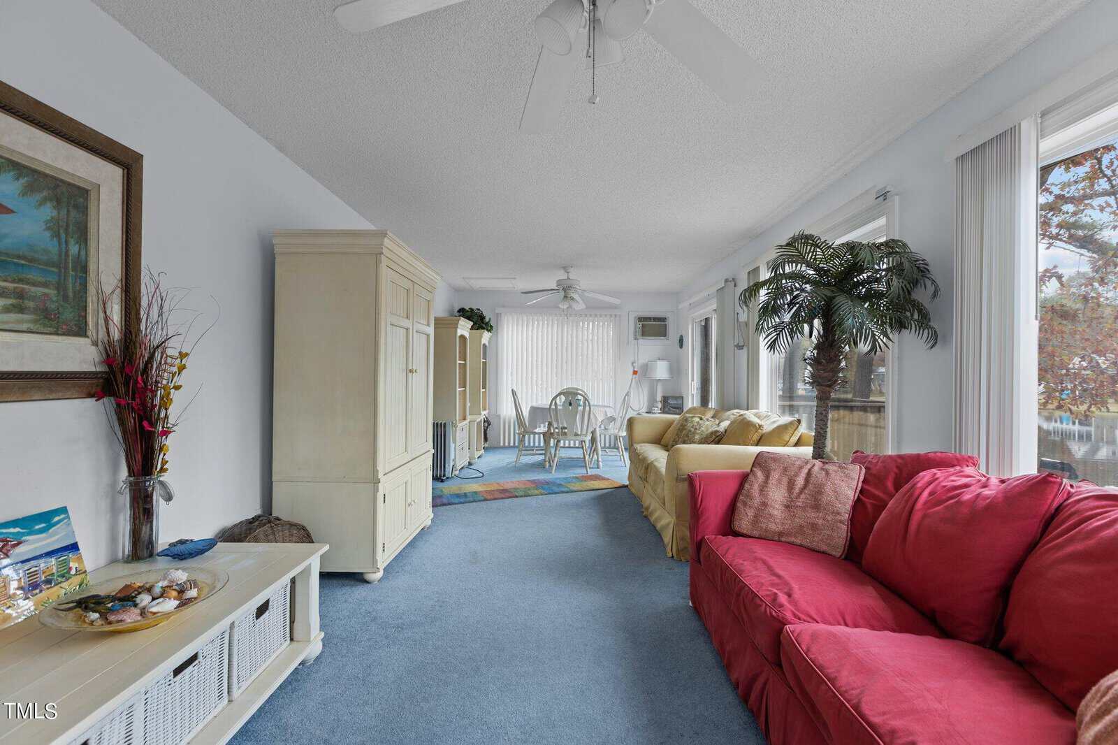A bright living room with blue carpet, a red sofa, yellow sofas, a white entertainment center, large windows, indoor plants, and a dining area with a round table and chairs in the background.