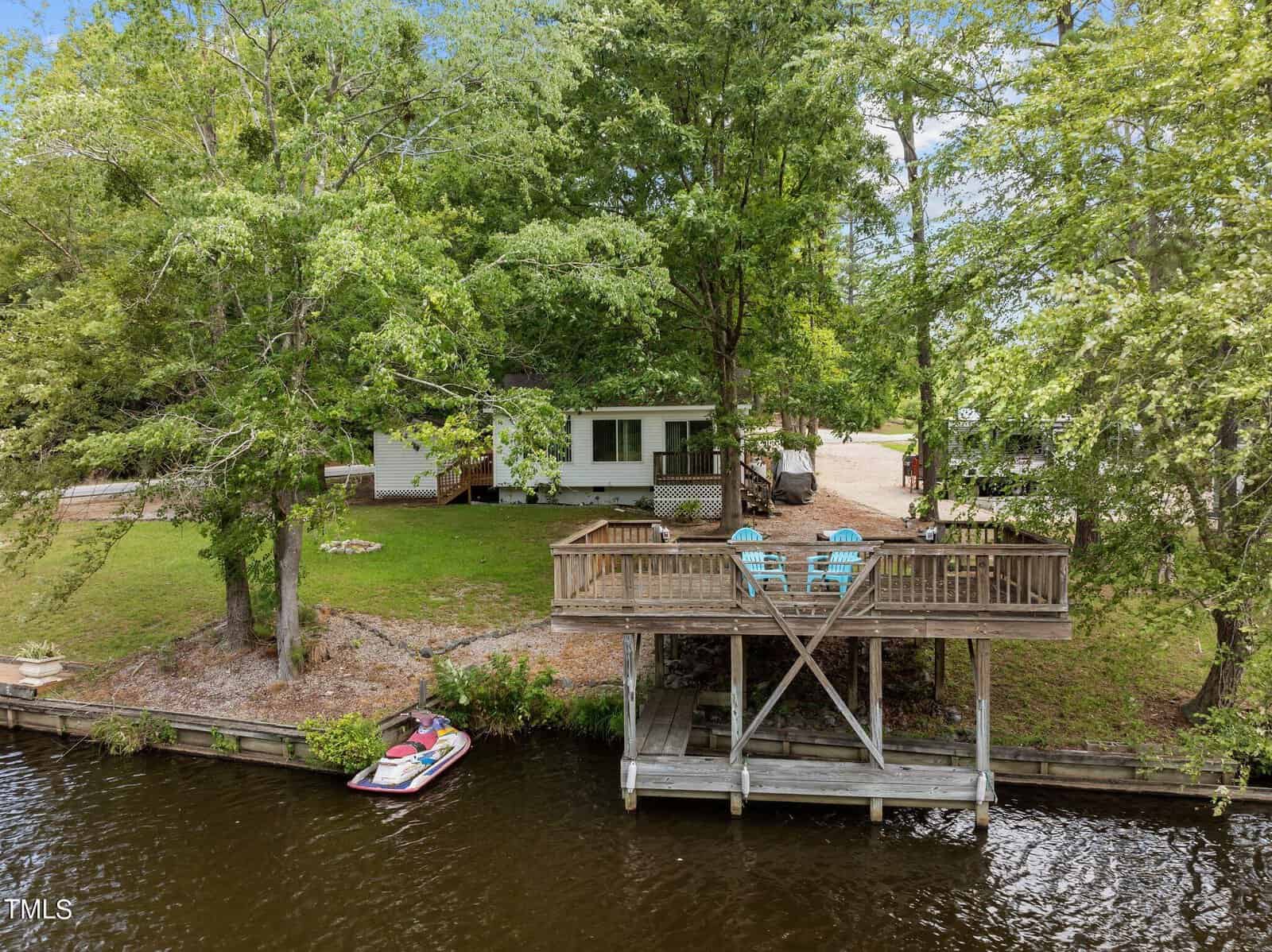 A small white house sits among trees beside a lake. In front, a wooden deck with blue chairs overlooks the water, and a jet ski is docked below the deck along the shoreline.