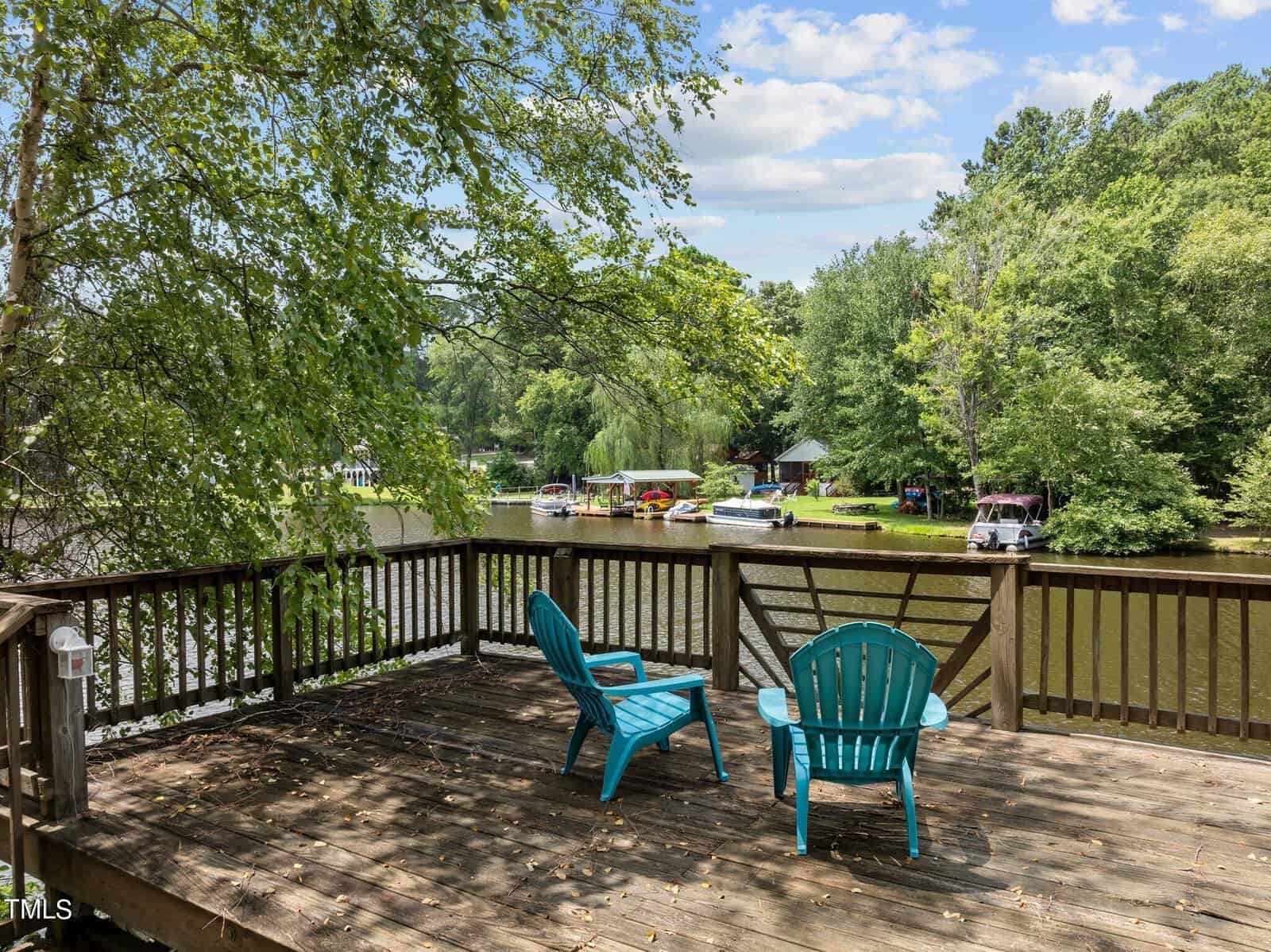 Two blue Adirondack chairs sit on a wooden deck overlooking a calm lake surrounded by trees, with several boats docked along the shoreline under a partly cloudy sky.