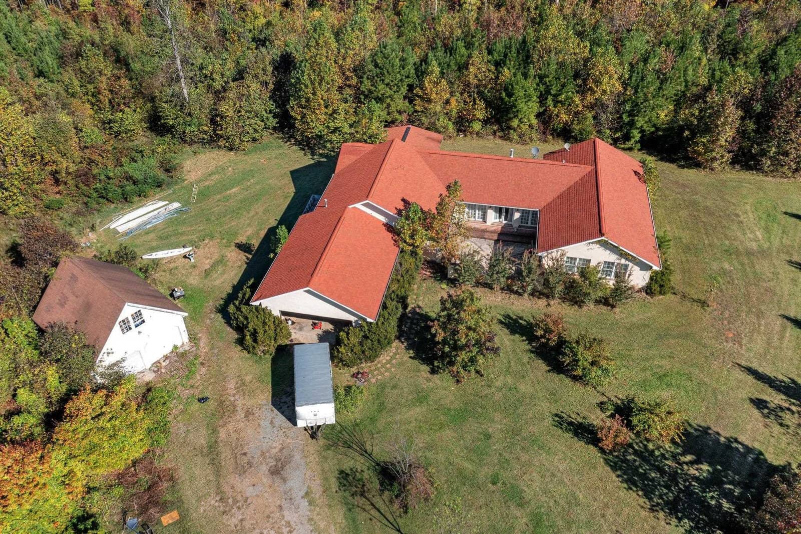 The aerial view showcases a large house with a red roof at 2701 Propus Wilkinson Road, surrounded by green trees. The property includes a small white building to the left and a tent-like structure adjacent to the main house. A grassy yard completes this serene Bullock setting.