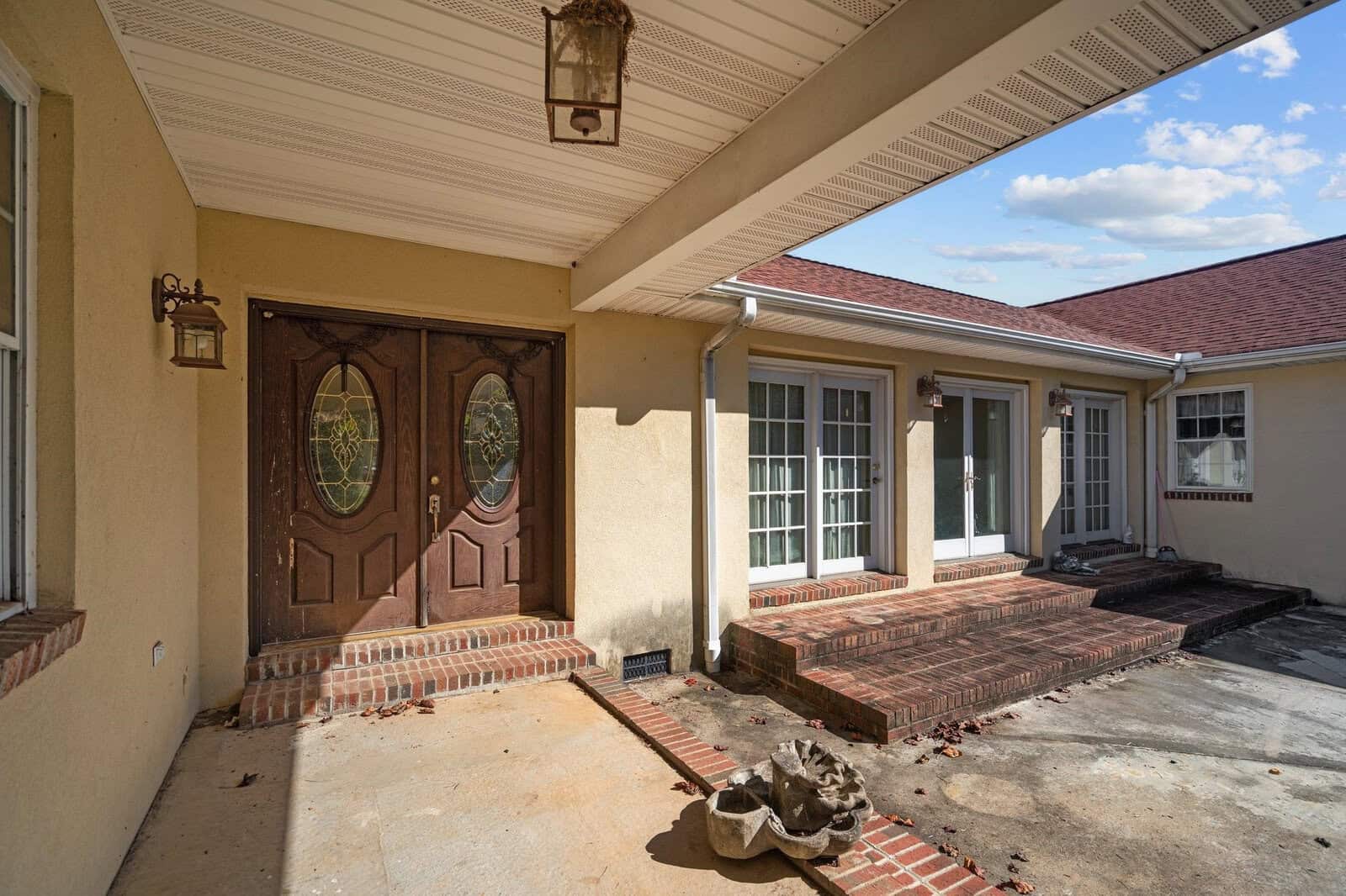 A beige house exterior with a covered porch featuring two wooden double doors and brick steps leading to a patio area. The porch has two hanging lantern-style lights. The sky is clear with a few clouds. A sculpture is placed on the concrete surface.