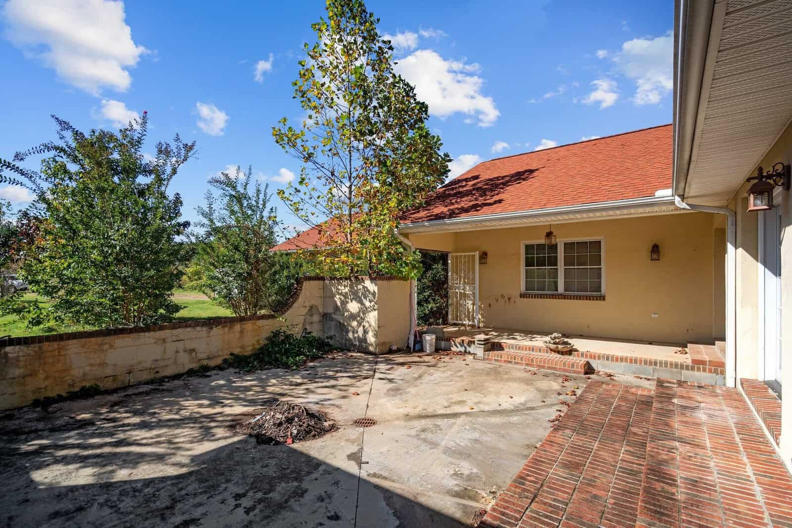 A small courtyard with a brick and concrete patio, featuring a tree and some plants. The house has a red roof and beige walls with a window and door. The sky is blue with a few clouds.