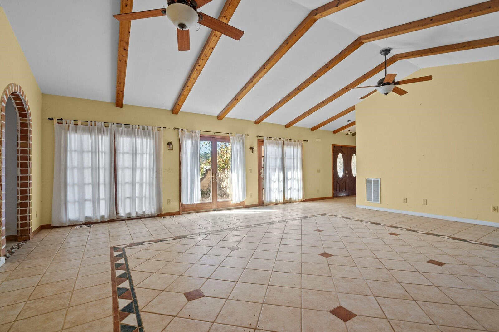 Spacious room with tiled floors, exposed wooden beams on the ceiling, and three ceiling fans. Large windows with sheer curtains let in natural light. There are arched brick doorways and a wooden front door.