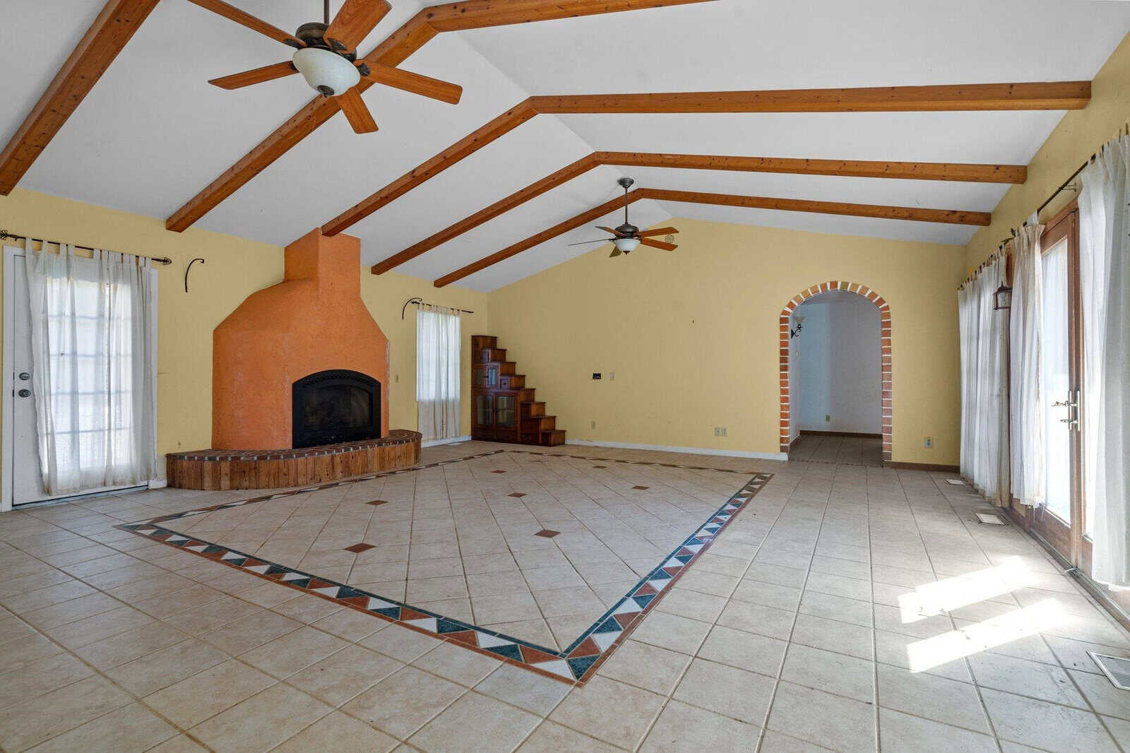 A spacious living room with a tiled floor, exposed wooden beams, and a large brick fireplace. Two ceiling fans hang from the ceiling. Sunlight streams through glass doors with curtains on both sides of the room. An arched doorway leads to another area.