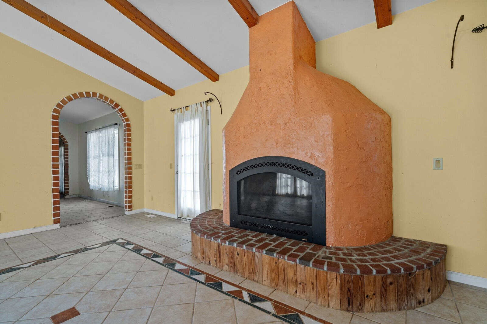 A room with a tall, orange stucco fireplace featuring a black metal frame. The floor is tiled with patterns, and wooden ceiling beams are visible. An arched doorway with brick trim leads to an adjacent space with sheer curtains on the windows.