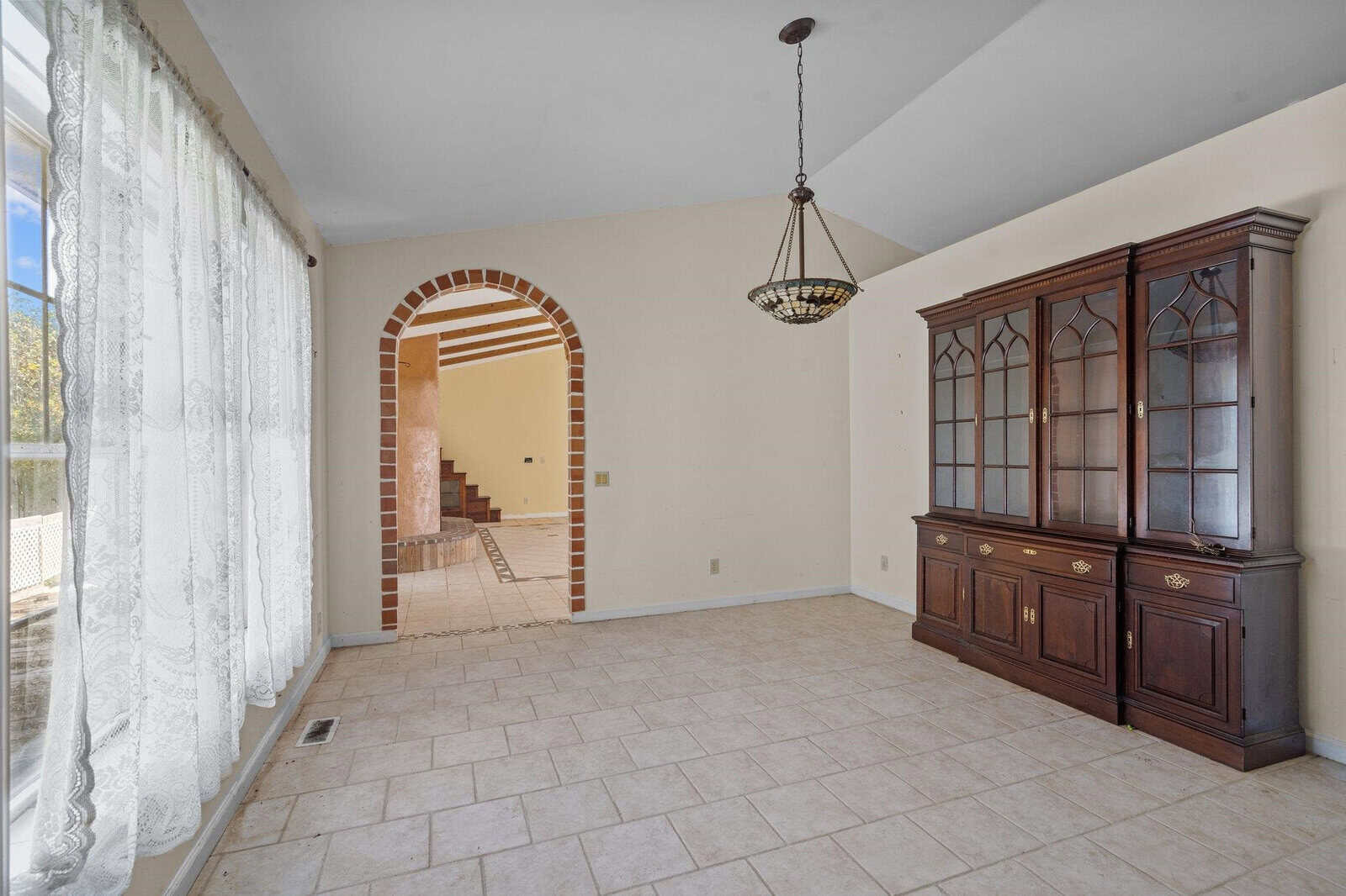 A bright, empty room with tiled flooring, featuring a large wooden cabinet with glass doors. A vintage chandelier hangs from the ceiling. An arched doorway leads to another area with wooden stairs and tile accents. Sheer curtains cover a large window.