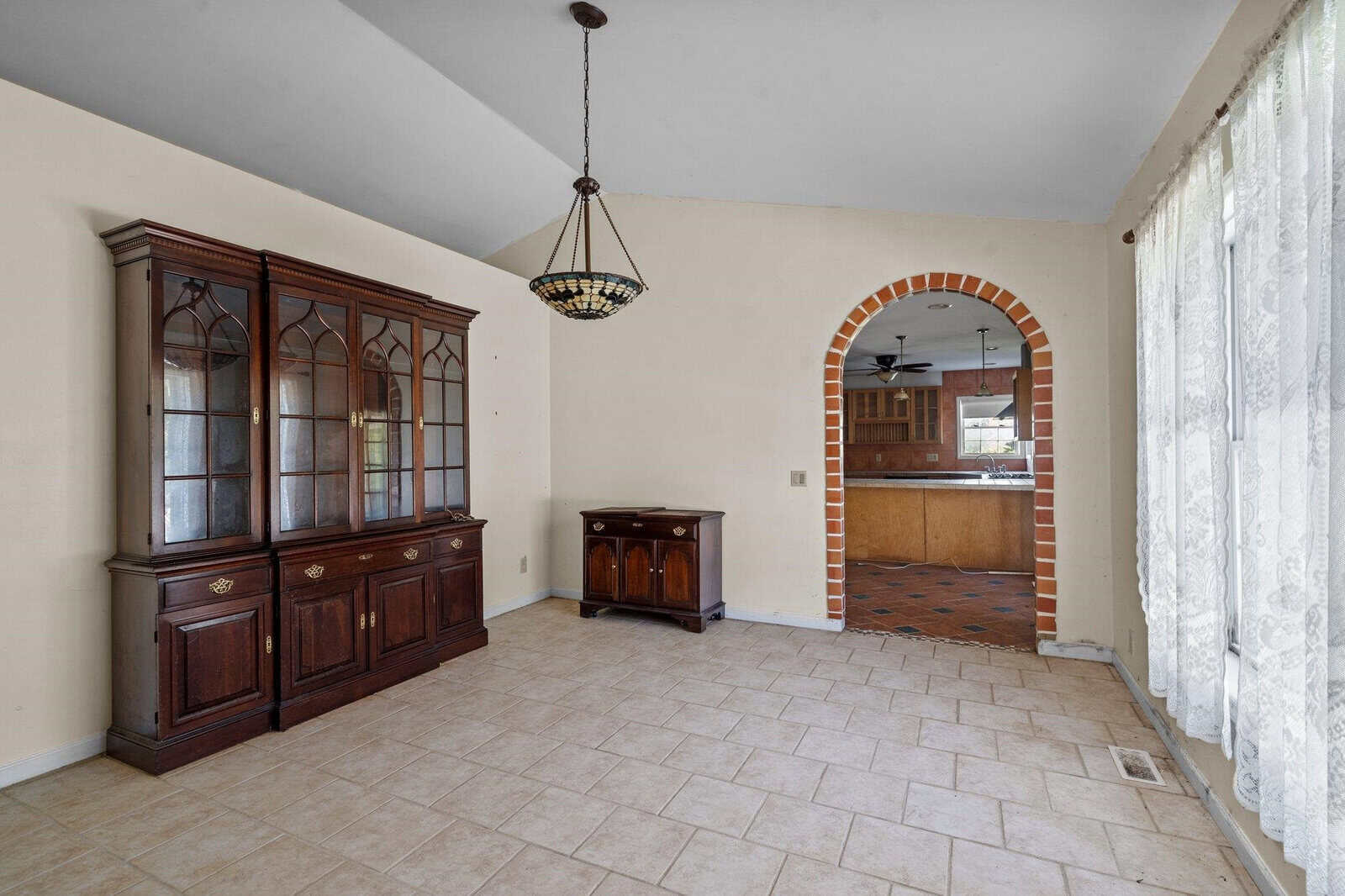 A room with beige tile flooring, a dark wooden cabinet on the left, and a matching chest underneath a vintage chandelier. There's a brick archway leading to a kitchen area with wooden cabinets and a ceiling fan. Lace curtains cover large windows on the right.