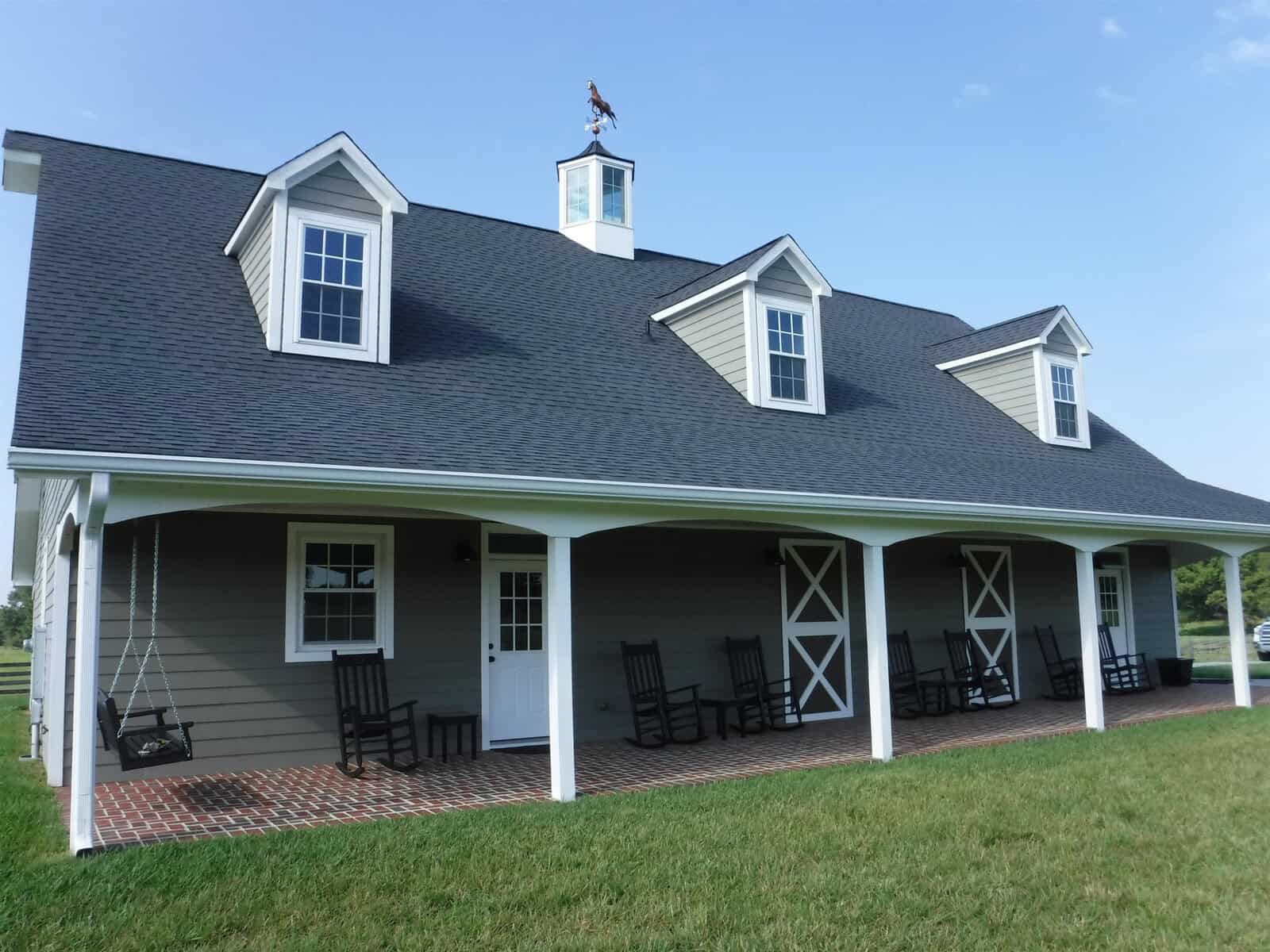 A large gray barn-style building with a steep black roof and three dormer windows. The front porch features multiple black rocking chairs and swings. A weather vane with a rooster adorns the rooftop, against a clear blue sky.