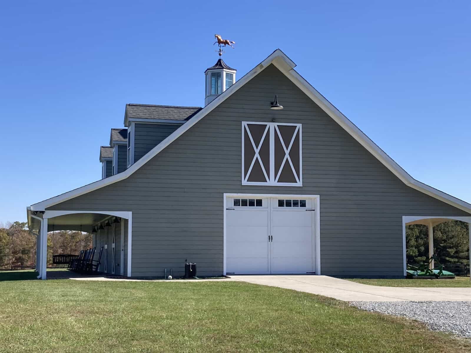 A modern barn with a gray exterior and white trim features double doors and a weather vane shaped like a horse on the roof. Surrounding the barn is a green lawn and clear blue sky.