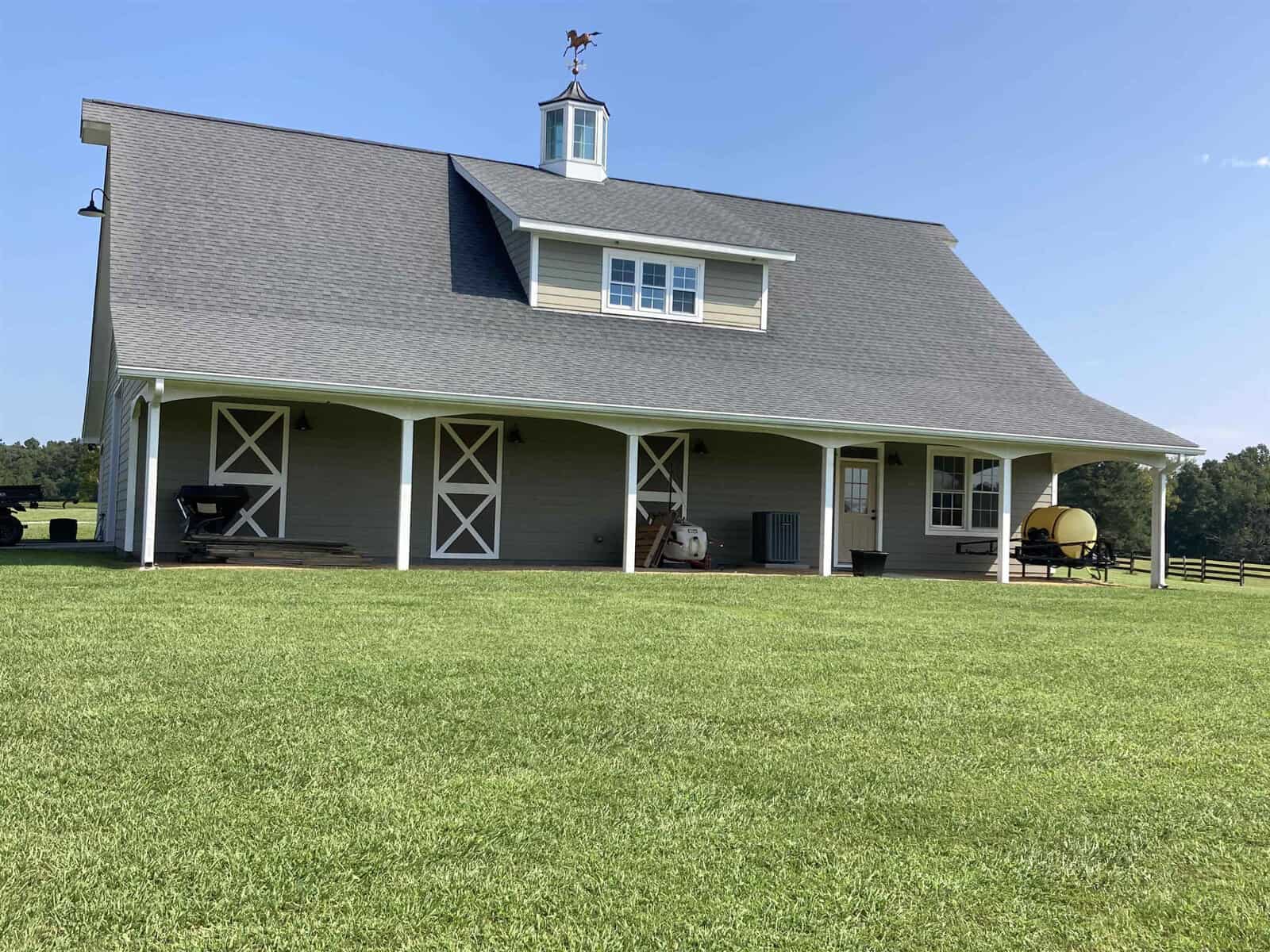 A large barn with a gray roof and light-colored siding sits on a well-manicured lawn under a clear blue sky. It features a small cupola with a weather vane. The barn has open storage areas and farm equipment nearby.