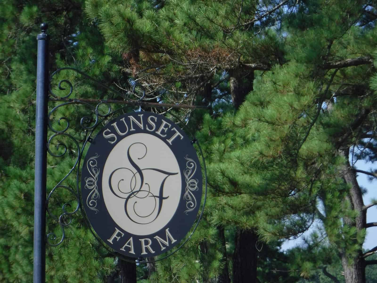 A round, black metal sign hangs against a backdrop of tall green pine trees. The sign reads "Sunset Farm" with the letters "SF" in the center, surrounded by decorative scrollwork.
