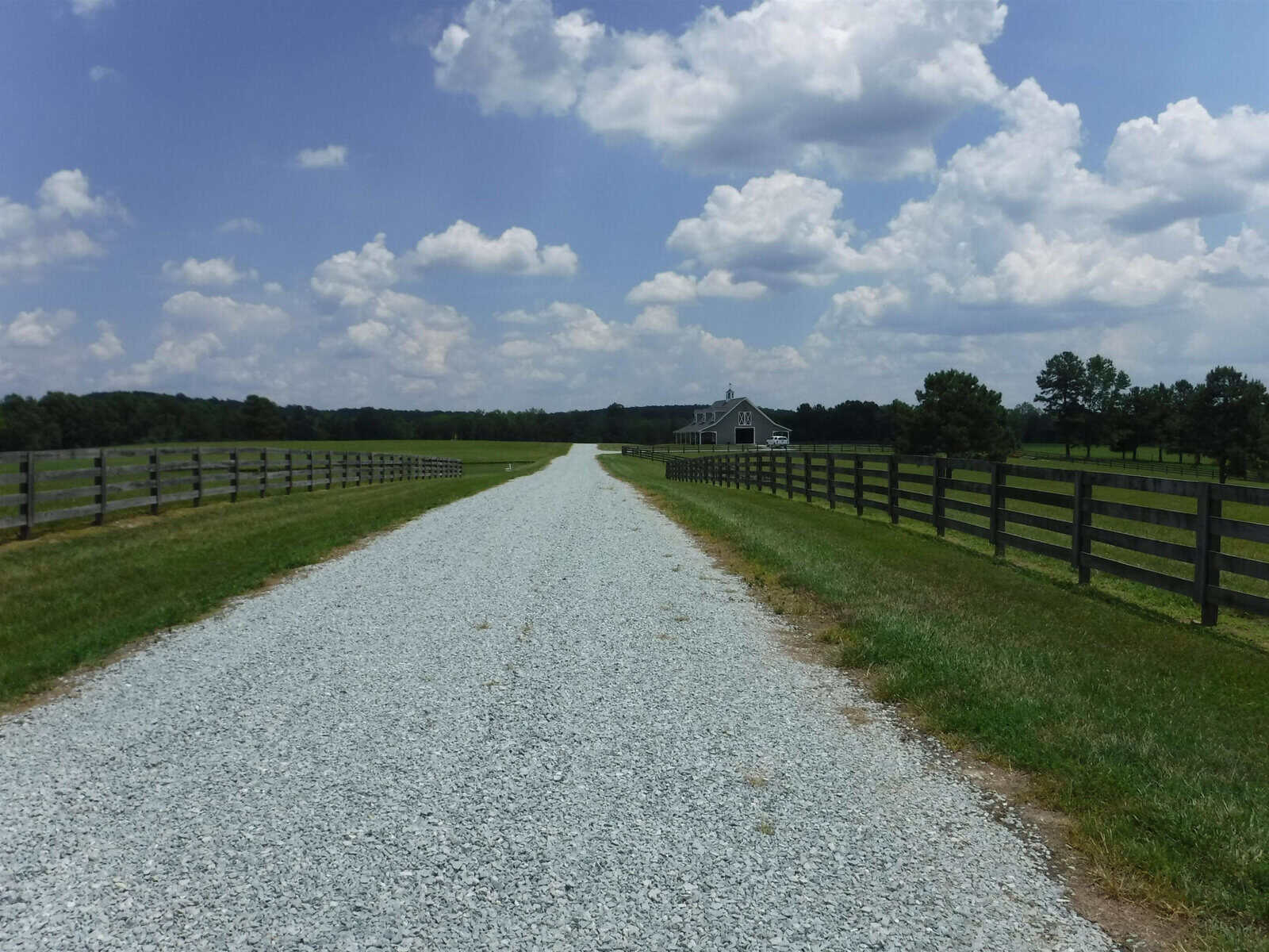 A gravel road leads to a distant barn surrounded by green fields and fenced pastures under a partly cloudy blue sky. Trees are visible on the horizon, adding to the serene rural landscape.